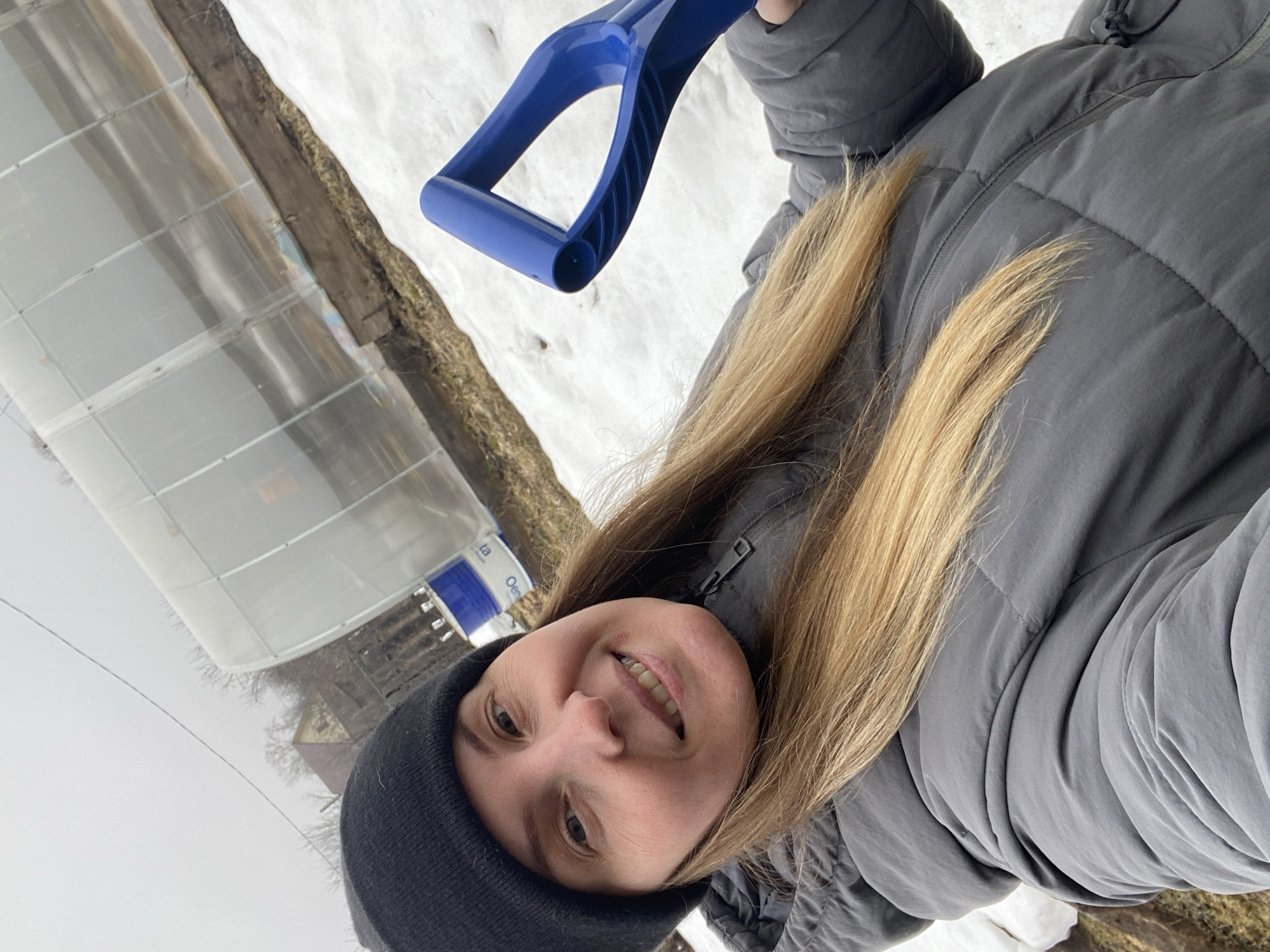 A woman wearing a black beanie and grey puffer jacket holds the blue handle of a small plastic shovel. She stands outside near a greenhouse with snow on the ground. The photograph may have been taken in Chaikovsky, Russia.