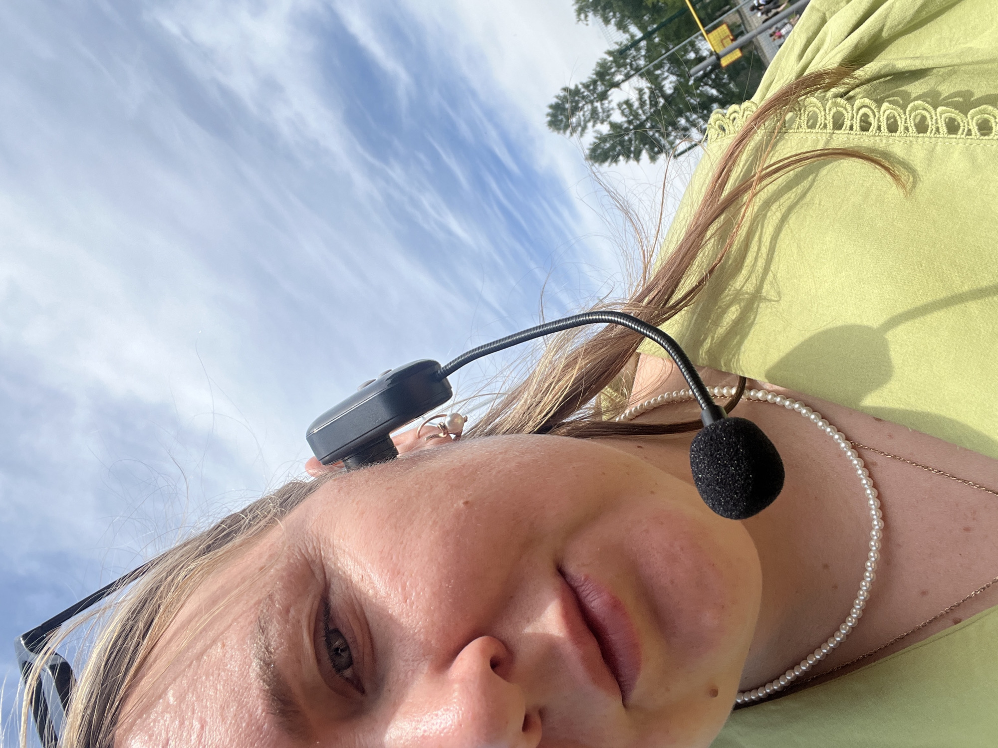 An extreme close-up, slightly Dutch-angled shot captures a person, likely female, in an outdoor setting in Chaikovsky, Russia. Her face dominates the foreground, tilted upwards towards a bright blue sky filled with wispy white clouds. She has light-colored hair and eyes, and a neutral, relaxed expression. A black headset with a flexible boom microphone is worn, with the microphone positioned near her mouth. Part of a dark-framed pair of glasses is visible near her left eye.
She is wearing a light green top with a decorative, scalloped lace or embroidered trim visible along the neckline. Adorning her neck are a strand of small white pearls and a delicate gold-colored chain. A small, light-colored ring is visible on one of her fingers.
In the background, against the clear sky, the tops of green trees are visible, suggesting a natural outdoor environment. Beyond the trees, hints of a structure with yellow elements, possibly a fence or part of a building like stadium seating, can be seen, indicating an event or sports venue. The bright sunlight casts soft shadows, suggesting it is daytime. The presence of the headset and microphone implies she might be engaged in an activity requiring hands-free communication, such as guiding, coaching, or participating in an outdoor event. No visible text is present in the image.