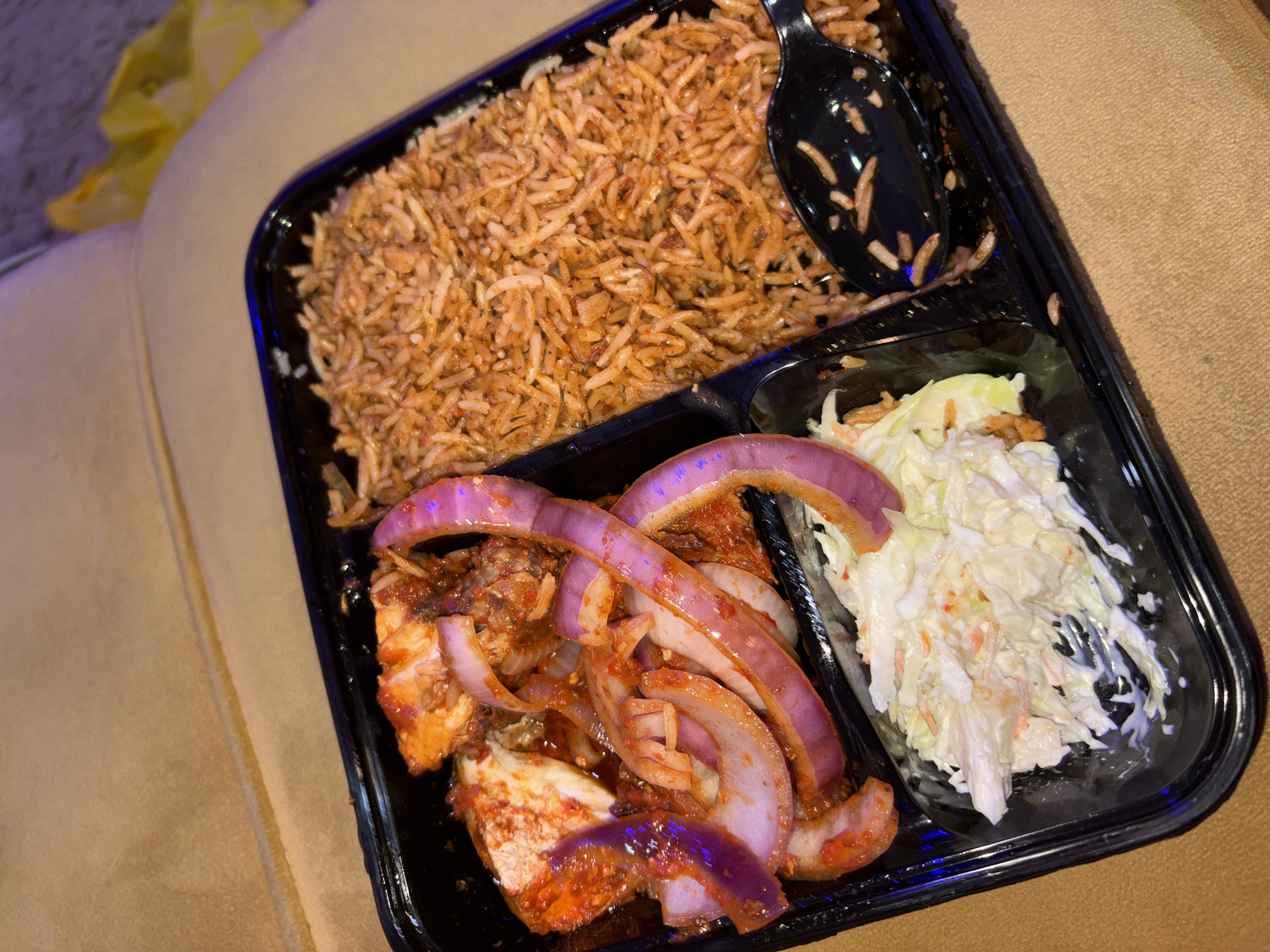 This media file shows a close-up, overhead view of a takeout container filled with food. The container is black plastic and appears to be sitting on a textured, beige surface, possibly upholstery.
The food itself is divided into three sections within the container. The largest section, at the top, contains a generous portion of rice. The rice is a reddish-brown color, suggesting it has been seasoned or cooked with a sauce, and appears to be long-grain. A black plastic spoon rests in this section of rice, with some grains clinging to it.
To the left and below the rice is a section with pieces of meat, likely chicken or fish, coated in a red sauce. Slices of red onion, with their characteristic purple hue, are layered on top of the meat.
The third section, to the right, contains a serving of coleslaw. The coleslaw is a mix of shredded white cabbage and carrots, bound with a creamy white dressing.
The lighting in the image suggests an indoor setting, likely in the evening or with artificial light. There are no people visible in the image, and no text is present. The overall impression is a prepared meal, ready to be eaten. Given the location context of Zanzi, Nigeria, this could be a typical meal from a local eatery.