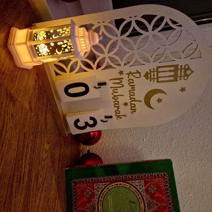 A Ramadan Mubarak decoration features a white cutout design with a lantern and crescent moon graphic. Beneath it, flip calendar blocks display the number "03." Above are two small red ornaments. A green and red book with ornate details sits below the ornaments. A decorative lantern with cut-out patterns is illuminated, resting next to the cutout design. The setting has a warm, inviting glow.
