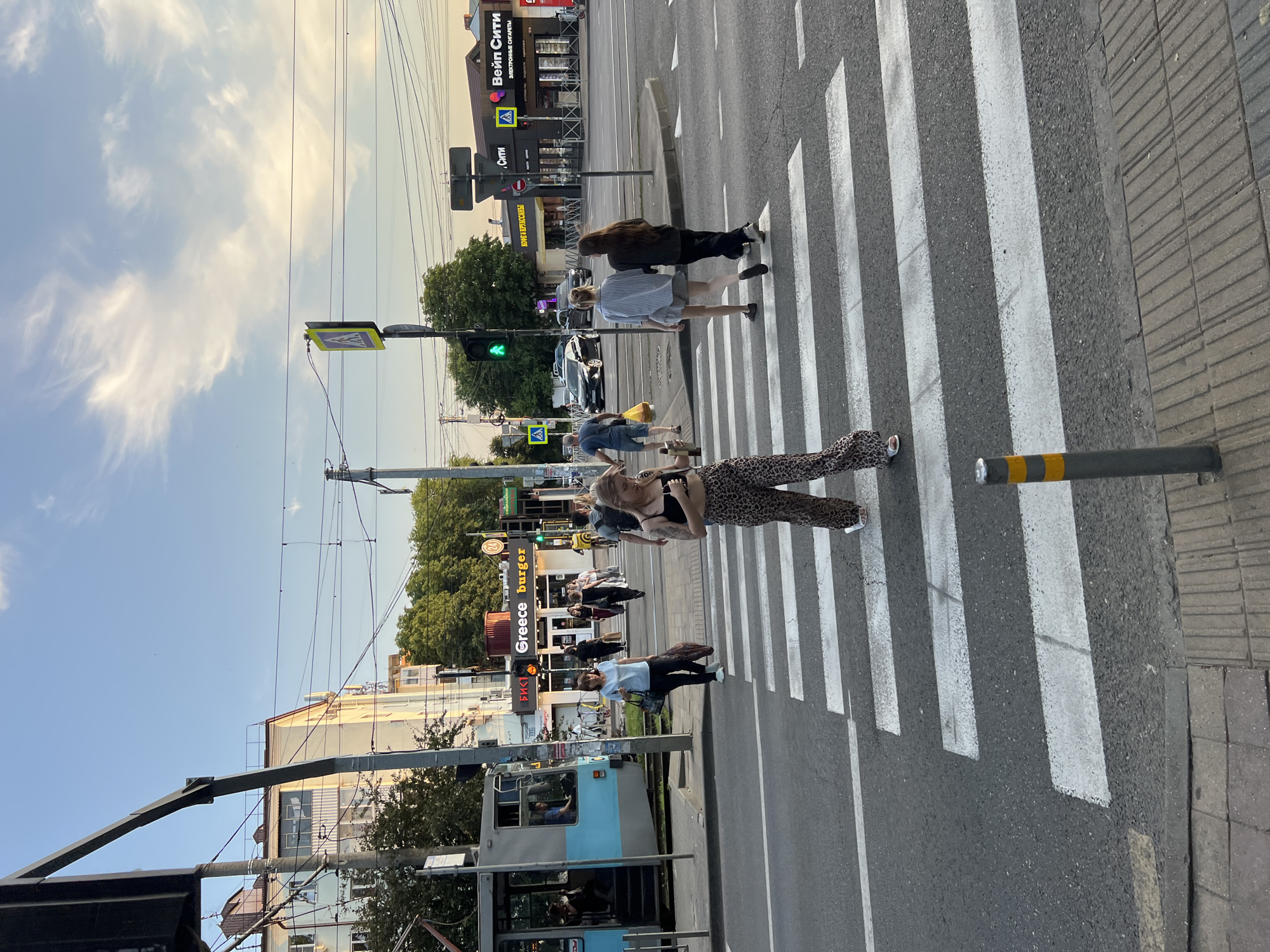 The image depicts a street scene in Krasnodar, Russia, likely during the daytime, with a clear blue sky and some wispy clouds. The perspective is from a low angle, looking up towards a pedestrian crossing. Several people are crossing the street, with a focus on a young woman in the foreground wearing a black top and leopard-print pants. She is looking towards the camera with a neutral expression. Behind her, other individuals are also crossing, some carrying bags, and one person is holding a yellow bag.
The scene includes a marked pedestrian crossing with white stripes on asphalt. On the left side of the street, a tram is visible, with passengers inside. Buildings line the street, including one with signage that reads "Greece Burger" and another with "ВЕЙП СИТИ" (Vape City) and "СИТИ" (City). Traffic lights are visible, with one showing a green light. Electrical wires crisscross above the street, suggesting an urban environment. The overall atmosphere appears to be that of a typical day in a city, with people going about their business. There are also bollards on the side of the road, one of which has yellow and grey markings. The image has been rotated and is presented in a vertical orientation.
