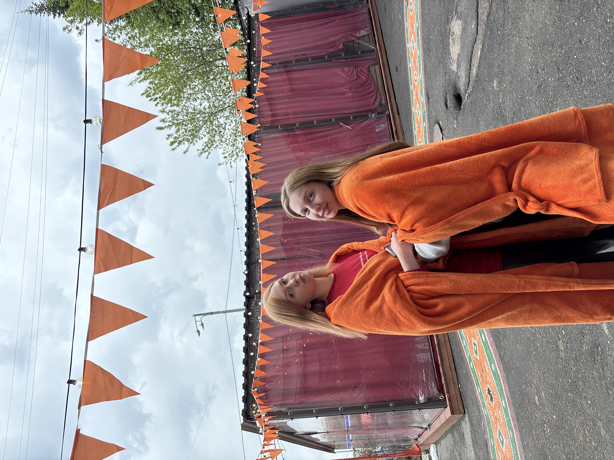 Two young girls stand next to each other, wrapped in orange blankets. They are outside a building decorated with orange triangle flags and clear plastic sheeting. The building has a maroon fabric backdrop. The ground next to the building has a decorative stripe with geometric patterns in orange, green, and white. The sky is overcast. Electrical wires and utility poles are visible in the background, along with trees. The location appears to be an outdoor area, possibly a street or sidewalk, in Perm, Russia.