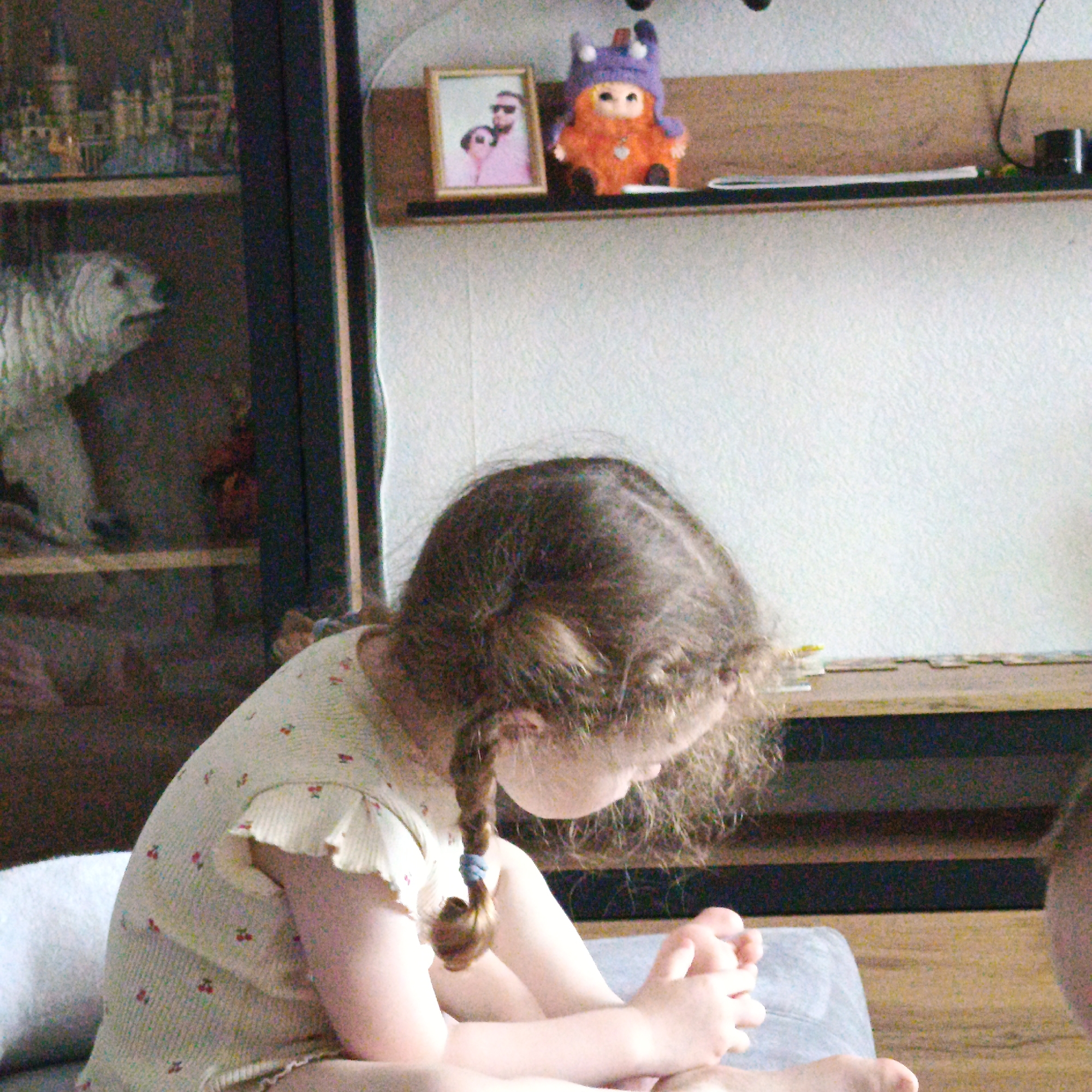 A young girl with brown hair in a braid sits on a gray cushion. Her head is bowed, and she is looking at her hands, which are clasped together. She is wearing a light-colored dress with cherry patterns and ruffled sleeves. In the background is a cabinet with a glass door containing various objects, including a white polar bear figurine. On a shelf above the cabinet is a framed photograph and a plush toy with a purple hat. A light-colored wall is visible behind the shelf. There is no verifiable evidence in the image to suggest that this scene is located in the city of Марковский, Russia.
