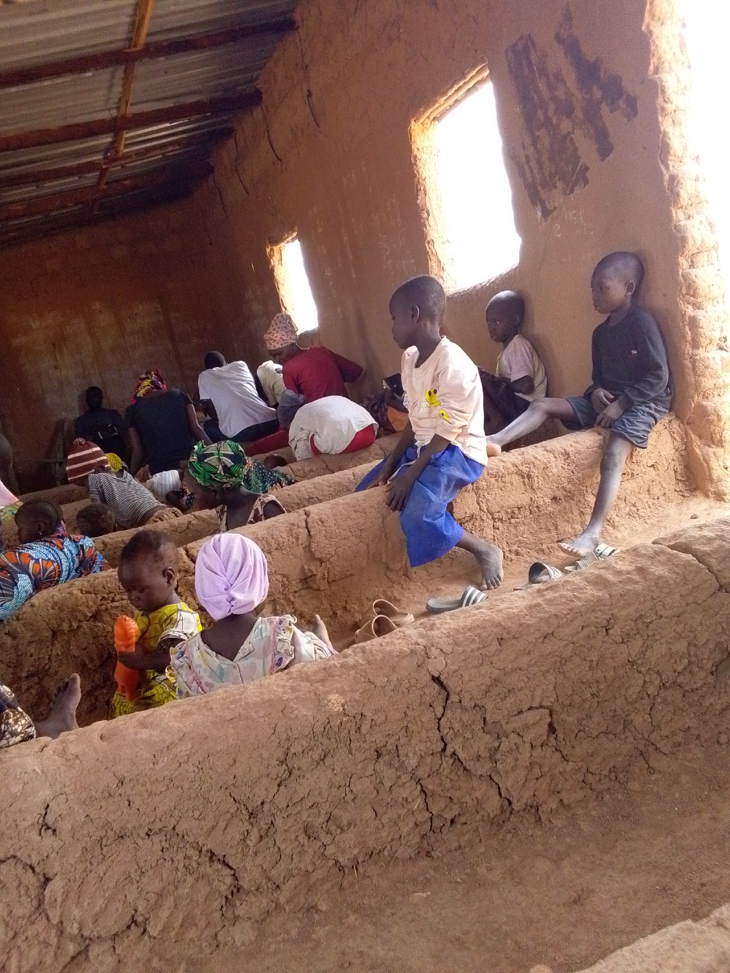 The image depicts a group of people, including children and adults, inside a building with earthen walls. The setting is likely in Mulke Barikin Sayejo, Nigeria, based on provided information. Individuals are seated on tiers formed from the earthen structure. Some are barefoot, and some have removed their sandals. Children are present in the foreground and middle ground. One child holds an orange object. Natural light streams through rectangular windows. The roof appears to be made of corrugated metal. Some individuals are lying down. Some wear head coverings.