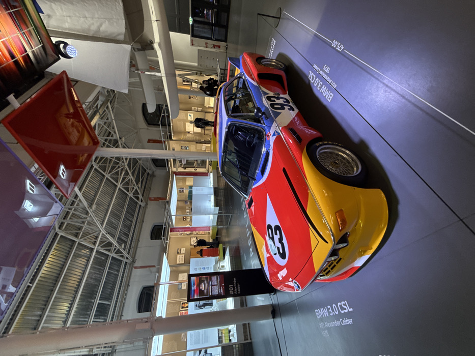 The image displays the vibrant BMW 3.0 CSL Art Car #01, designed by Alexander Calder in 1975, presented in an indoor museum or exhibition hall, likely located in Milan, Italy. The car is the primary subject, featuring a distinctive paint scheme of bold red, yellow, blue, and white sections with black accents, and the race number "93" visible on its side.
The car is positioned on a dark gray floor that has white lines and text indicating "BMW 3.0 CSL," "#01 Alexander Calder," and "1975." Another marking, "475.00-", is also visible on the floor. In the background, the scene reveals a spacious hall with high ceilings and exposed white metal trusswork, suggesting an industrial or contemporary architectural style. Various other exhibits and display cases, some illuminated, line the walls and floor, creating a gallery-like atmosphere. A digital display screen in the mid-ground shows the text "REPLAY." A few indistinct figures, appearing to be museum visitors, can be seen observing other parts of the exhibition. The lighting is indirect, highlighting the car while maintaining a subdued ambiance in the background. The event captured is a public display of an iconic art car within a museum context.