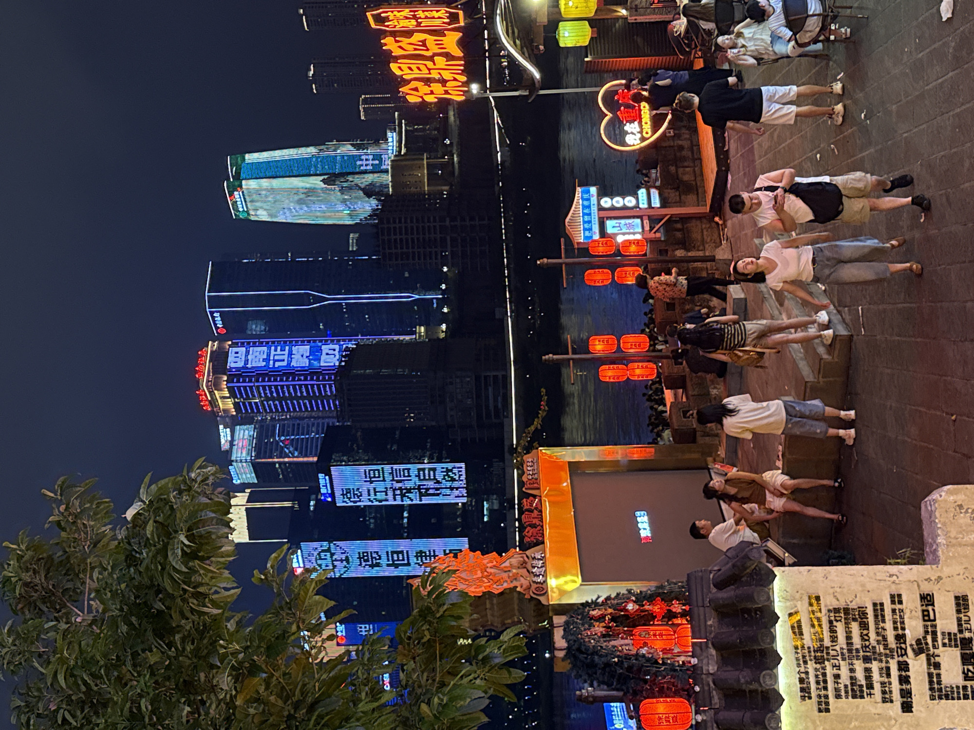 This image captures a nighttime scene along a waterfront promenade, likely in the Yuzhong District of Chongqing, China. Tall, illuminated skyscrapers dominate the background, their facades covered in vibrant LED displays showcasing Chinese characters and geometric patterns. In the foreground, a group of people are seen walking and sitting, enjoying the evening atmosphere. Red lanterns are strung along a railing, adding a festive touch. A large, bright yellow sign with red Chinese characters is prominently displayed, suggesting a commercial establishment. A body of water is visible between the promenade and the city skyline, reflecting the colorful lights. The ground is paved with stone, and some of the people are dressed in casual attire, indicative of a relaxed urban environment.