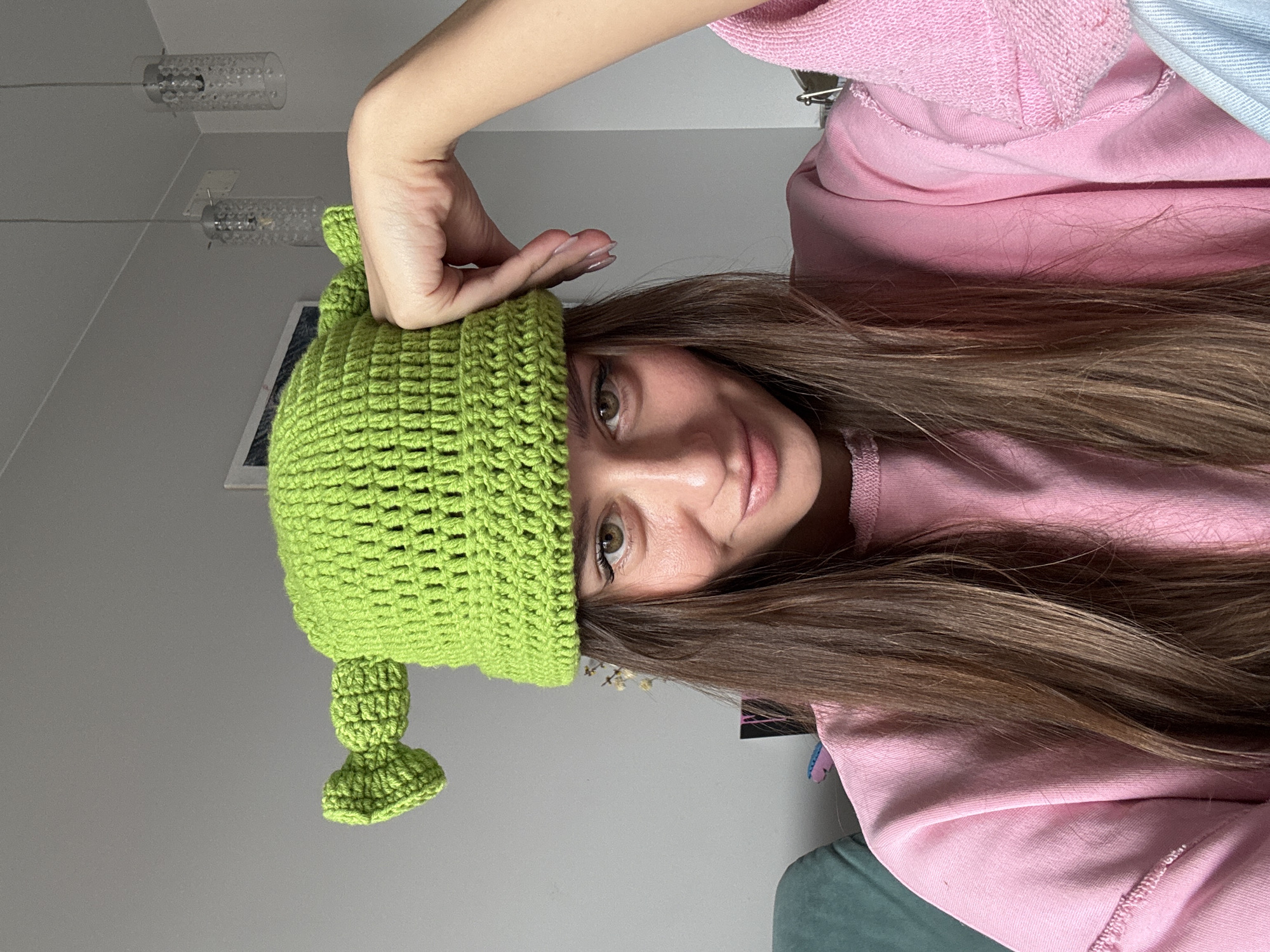 A young woman is captured in a close-up, head-and-shoulders portrait, likely taken indoors in Krasnoyarsk, Russia. She looks directly at the camera with a soft, inviting gaze and a subtle smile. Her long, straight dark brown hair cascades around her shoulders.
She is wearing a distinctive, bright green crocheted hat designed with ear-like protrusions, reminiscent of the character Shrek. Her left hand is gently placed on the brim of the hat. She is dressed in a soft pink hooded sweatshirt, which covers her upper body.
The background is simple, featuring a plain light gray wall and ceiling. Two modern, cylindrical light fixtures, composed of numerous small, clear beads, are visible suspended from the ceiling. A faint, partially obscured outline of what appears to be a framed picture can be seen on the wall behind her. The lighting is even and soft, suggesting an indoor setting under artificial light. There is no visible text in the image.