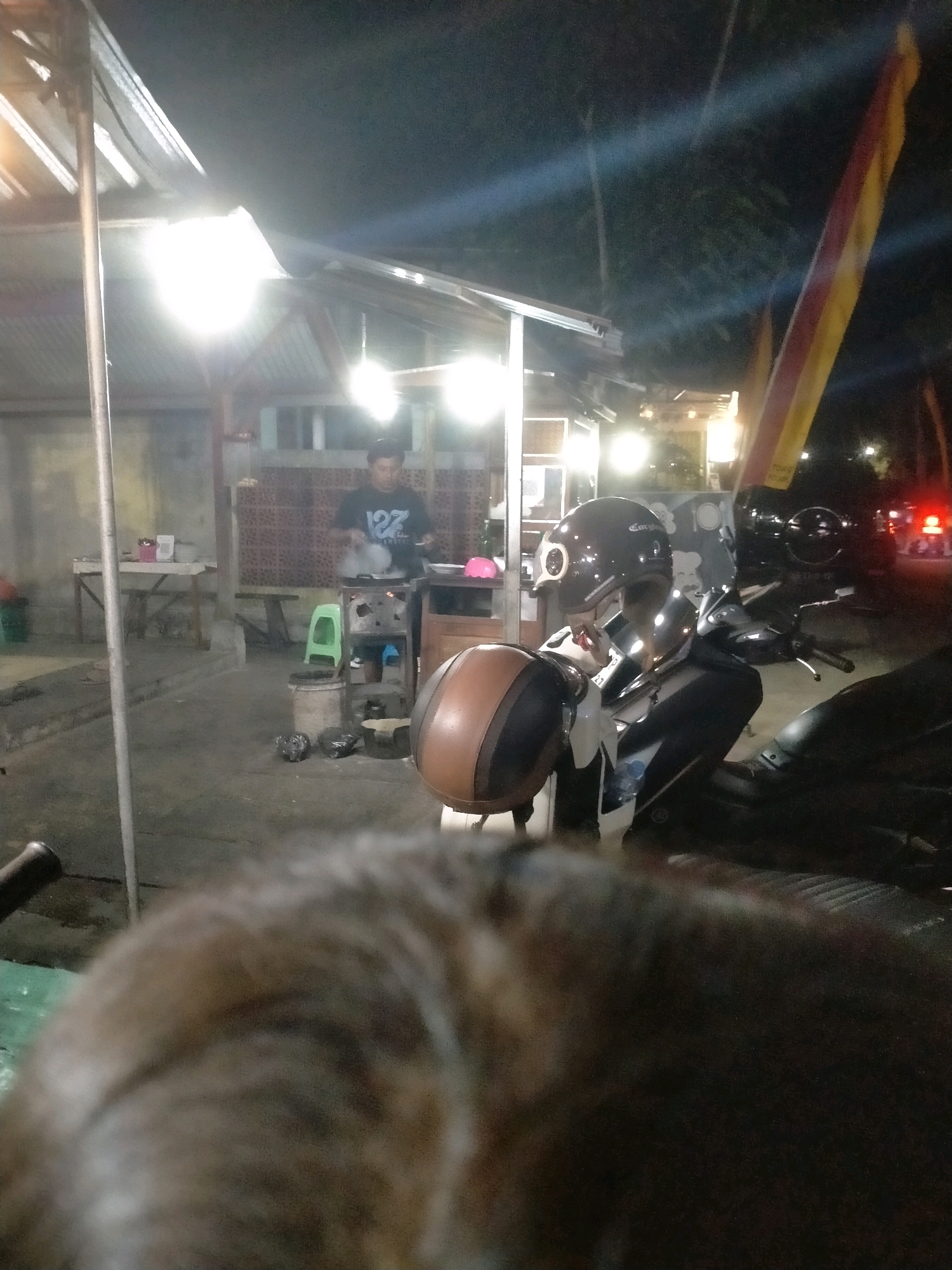 The image depicts an outdoor night scene in what appears to be a food stall or small eatery in Playen, Indonesia. A man in a dark t-shirt with a white graphic is standing and cooking at a portable stove, with steam rising from a pan. He is positioned under an awning illuminated by bright lights. In the foreground, to the right, a dark-colored motorcycle is parked, with a helmet placed on its handlebars. Another helmet, brown and black, is resting on a different surface to the left of the motorcycle. Various objects, including a green plastic stool, buckets, and what appear to be bags of trash, are scattered around the area. The background shows a building with a tiled facade and more lighting. A flag with red and yellow stripes is visible on the right side of the frame. The image is taken from a low angle, with the foreground largely out of focus, suggesting the camera is positioned close to a furry object, possibly an animal.