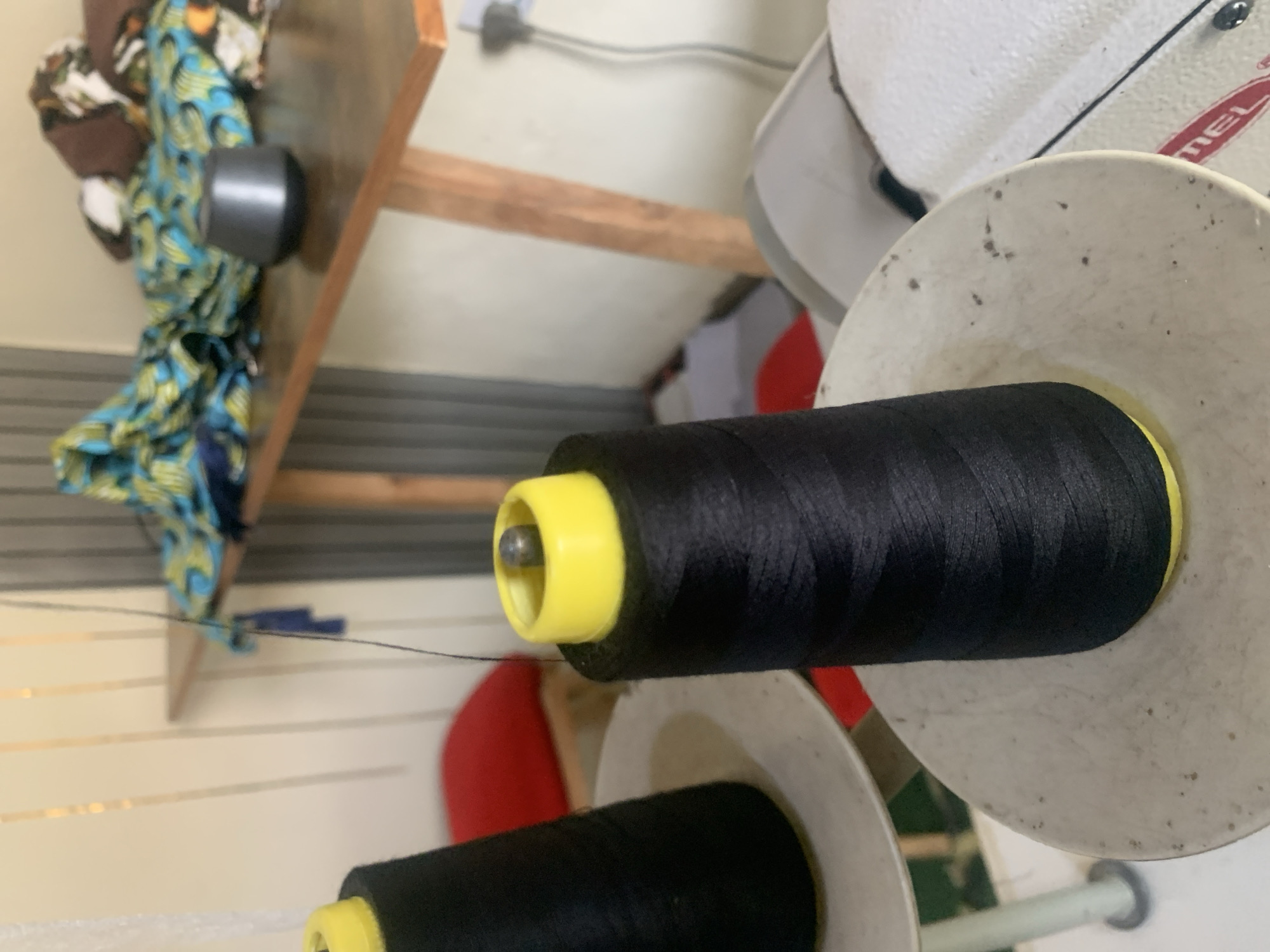 The image shows a close-up of two spools of black thread, one in the foreground and another slightly behind it to the left. Both spools have bright yellow caps, and the thread is tightly wound. The spools appear to be part of sewing equipment, likely industrial sewing machines, as suggested by the white housing of a machine visible on the right side of the frame. In the background, a wooden table holds several pieces of colorful fabric with a blue and green pattern, along with a small, dark gray, dome-shaped object. The wall behind the table is paneled with vertical elements. The scene is lit by artificial light, casting soft shadows. The overall impression is that of a workshop or tailoring area. The location of Jauro Yinu, Nigeria, cannot be determined from the visual information provided in the image.
