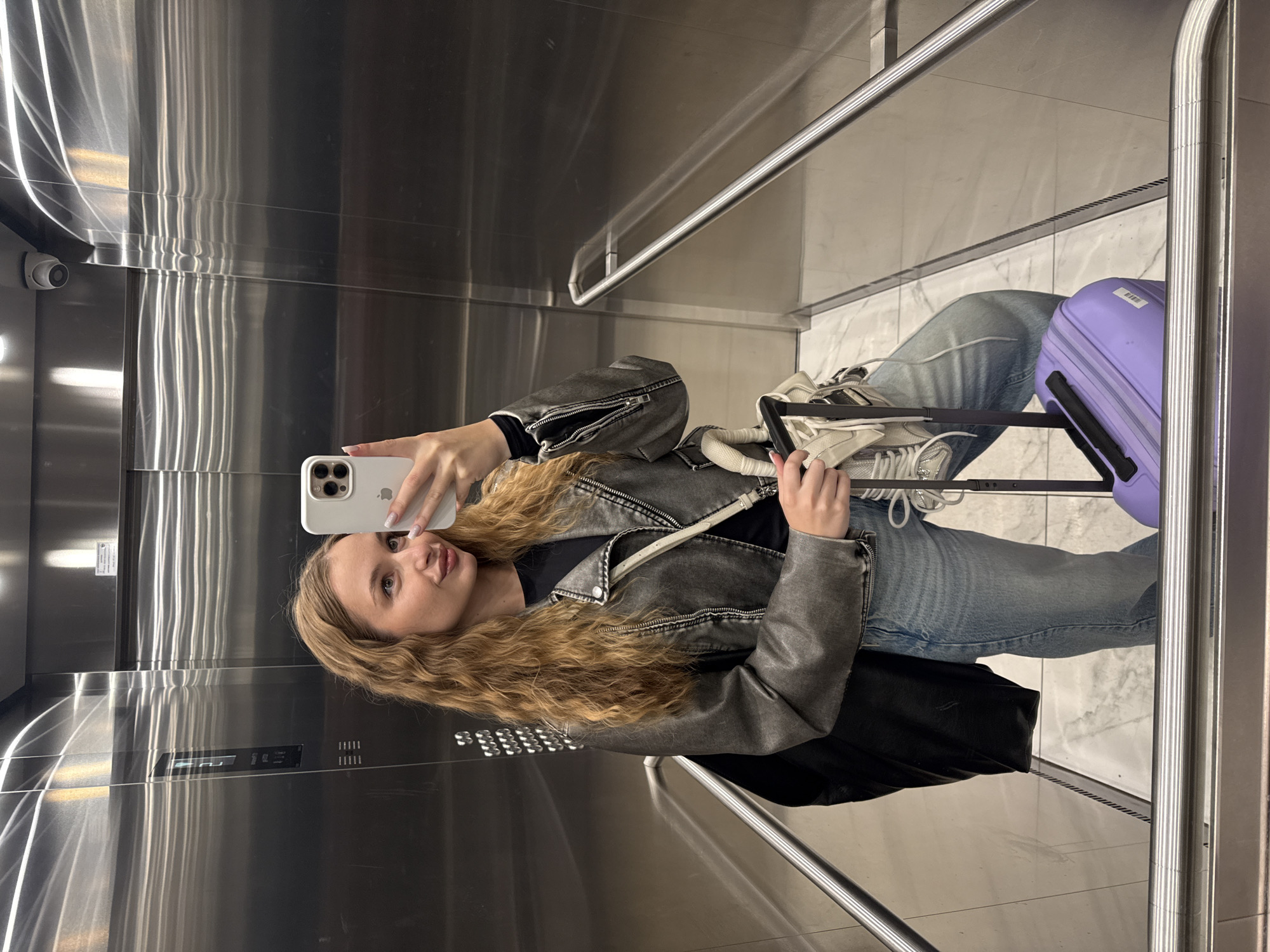 A young woman is taking a selfie in an elevator with shiny metal walls, possibly in Belgrade, Serbia. She is holding a white smartphone in her left hand and is wearing a gray jacket, black shirt, and blue jeans. She has long, wavy blonde hair. She is standing near the elevator door, and behind her is a purple suitcase and a pair of white sneakers placed atop the suitcase handle. The elevator's interior has stainless steel walls and a marble-patterned floor. A security camera is visible in the top left corner of the elevator.