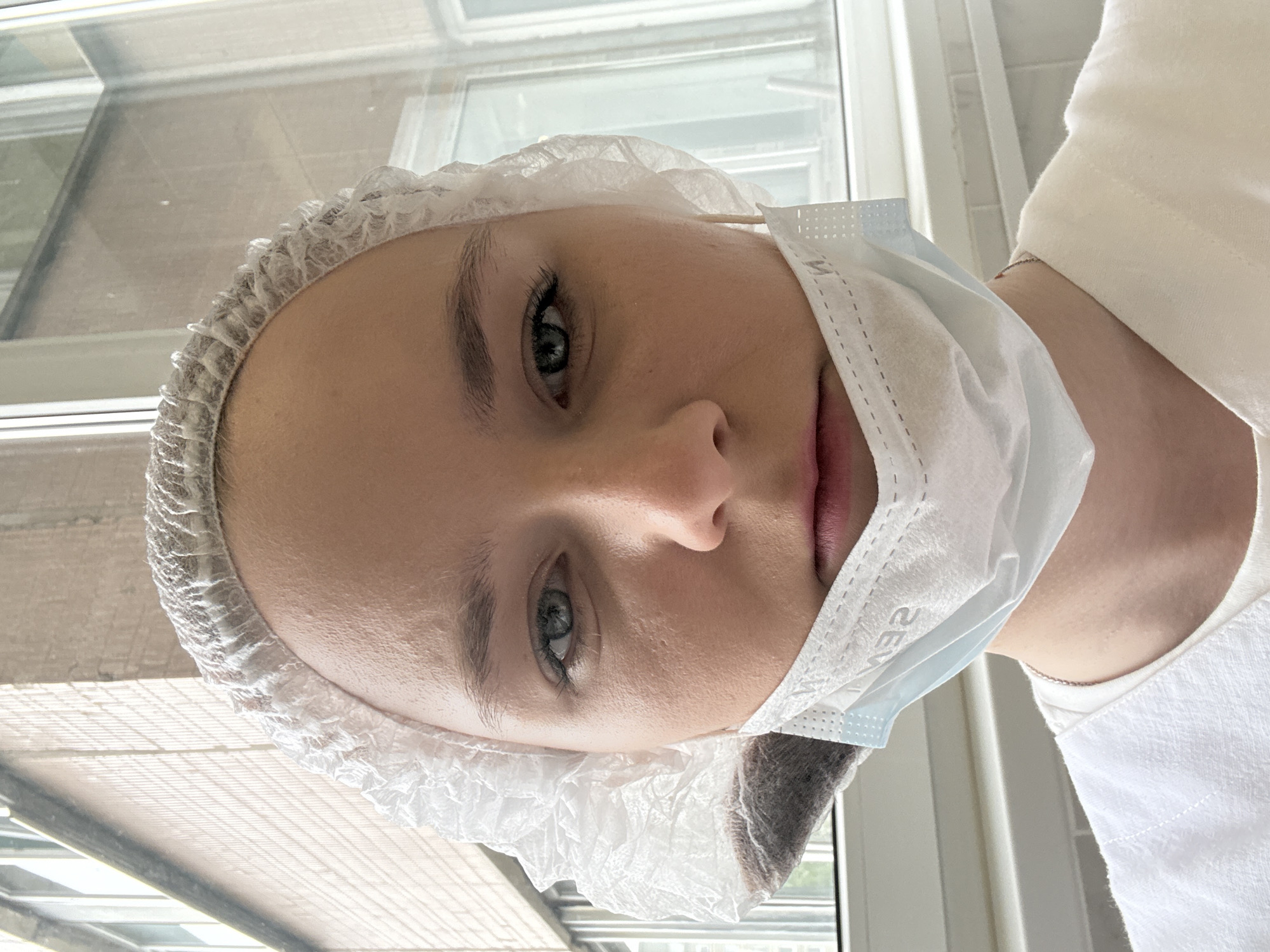A young woman is shown in a close-up shot, wearing a white disposable hairnet and a light blue disposable face mask pulled down to her chin. Her eyes are gray-blue, and she appears to be wearing minimal makeup. The background shows a window with a grid pattern, indicating an institutional setting. The location is assumed to be in Saint Petersburg, Russia. The subject is wearing a white top, likely a uniform. The image suggests a healthcare or clinical environment, possibly a hospital or beauty salon.
