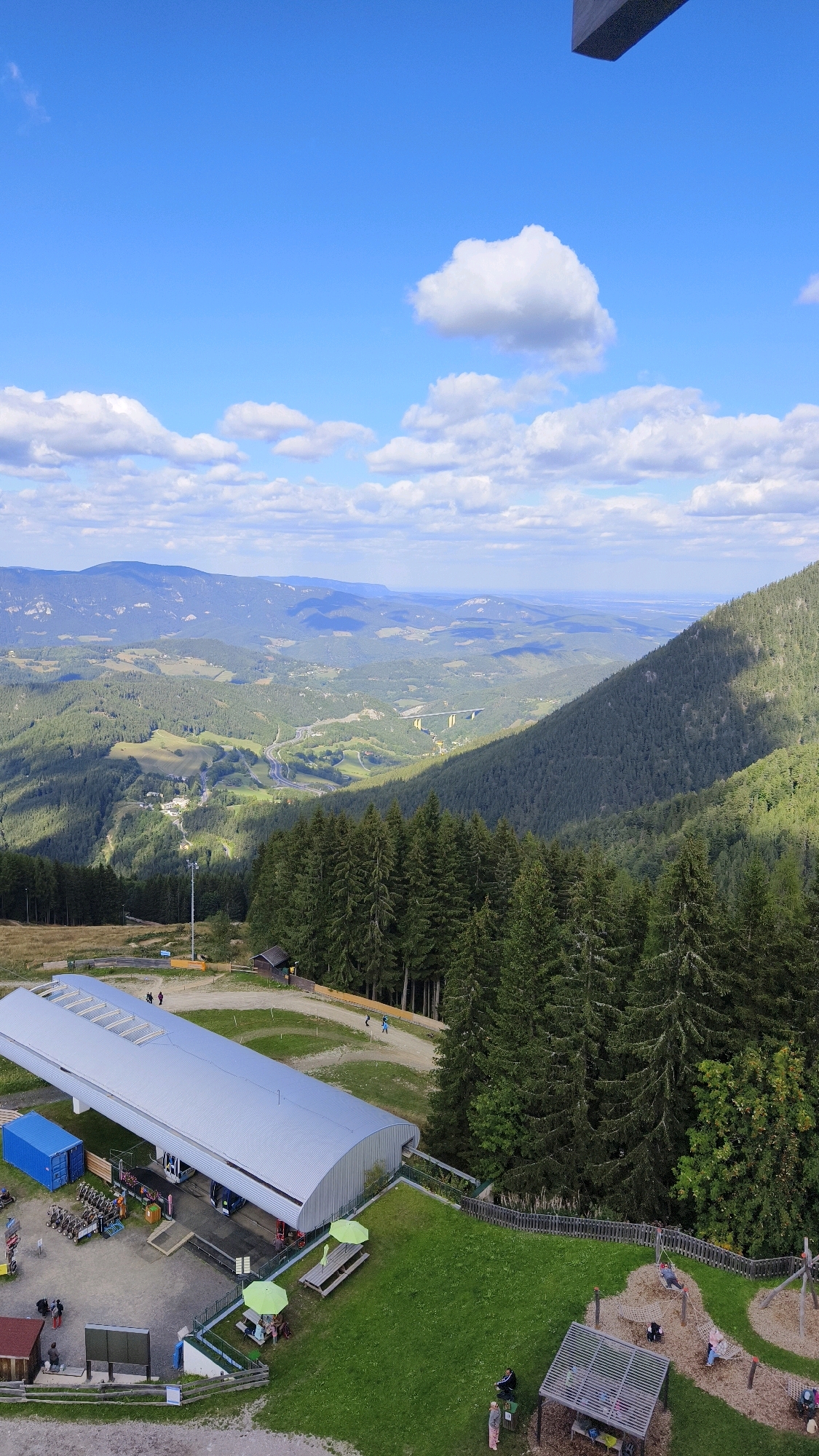 This image captures a panoramic view from a mountain in Semmering, Austria, likely taken from an elevated vantage point, possibly a viewing platform or a mountain station. The scene is dominated by a vast landscape of rolling hills covered in dense evergreen forests, indicative of the alpine region.
In the foreground, a modern building with a silver, corrugated metal roof is visible. This structure appears to be a station or a facility, possibly for a cable car or a ski lift, given the presence of what looks like a track extending from it. Adjacent to the building, there's a blue shipping container. Several vehicles, resembling ATVs or small utility vehicles, are parked near the station, along with some equipment.
To the right of the building, a grassy area features two picnic tables with bright green umbrellas, and a children's playground with swings and climbing structures. Some people are present in this area, engaged in leisure activities.
The mid-ground reveals a valley with a winding highway and a prominent yellow bridge spanning across it, suggesting a significant infrastructure in the area. The valley is dotted with scattered buildings and patches of cleared land, interspersed with more forested hills.
The background consists of multiple layers of mountains receding into the distance, fading into a hazy blue. The sky above is a clear blue, punctuated by scattered fluffy white clouds, suggesting fair weather. The overall impression is of a scenic mountain resort area, offering both natural beauty and recreational facilities.