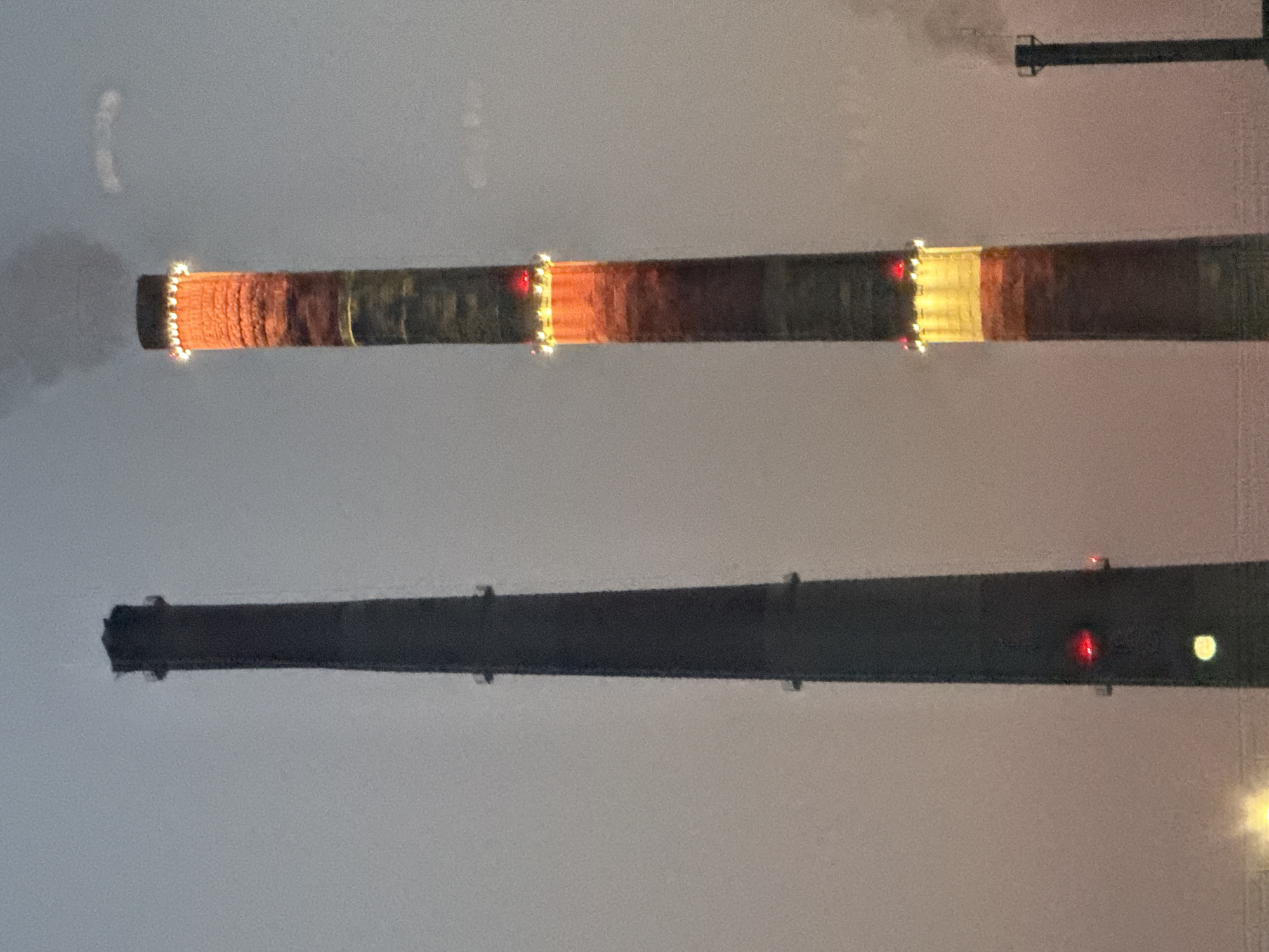 The image shows two industrial smokestacks against a hazy, muted sky, likely during twilight or dawn. The smokestack on the left is dark gray and appears solid, with two red lights and one yellow light visible on its side. The smokestack on the right is taller and emits a plume of dark smoke. It is adorned with a ring of bright lights near its top, which cast an orange glow on the upper section of the stack. This stack also has bands of reddish-brown and gray. A smaller, darker smokestack is visible in the far right background, also emitting smoke. The overall scene suggests an industrial area, consistent with the heavy industry present in Norilsk, Russia. There are no people or animals visible in the frame.