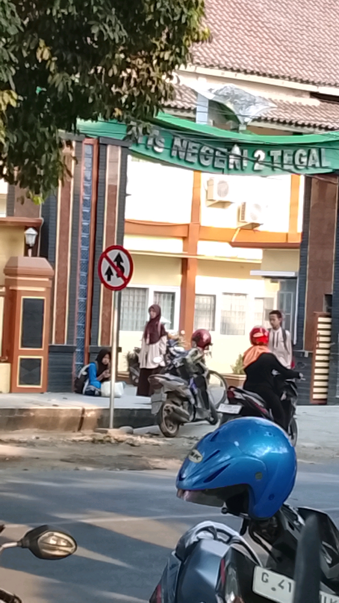 The image captures a street scene in front of a building identified by a green banner reading "IIS NEGERI 2 TEGAL". The building appears to be a school or government institution with light-colored walls and windows. A tree with dark green foliage hangs over the left side of the frame. In the foreground, several motorcycles are parked, with a prominent blue helmet placed on one. On the sidewalk in front of the building, two young women are seated, one wearing a blue jacket and the other in a brown hijab and dress. Another young woman, also in a hijab, stands near a motorcycle, and further back, a young man in a light-colored shirt is visible. A traffic sign with a red circle and a diagonal line, indicating a prohibition against two-way traffic, is positioned near the seated individuals. The street is paved and appears to be in moderate sunlight. The overall atmosphere suggests a typical day in a populated area.