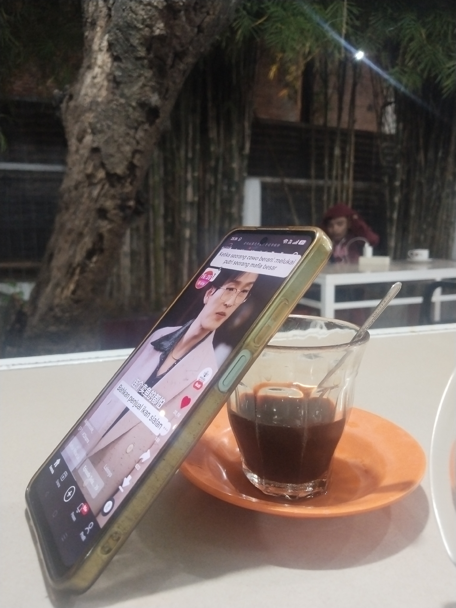 The image captures a scene at a table, likely in a cafe or outdoor seating area in Lhokseumawe, Indonesia. A smartphone is leaning against a glass cup filled with a dark beverage (likely coffee) on a small orange saucer. The screen displays a video, showing a man with glasses wearing a blazer. The video is likely from a streaming platform like TikTok or a similar service, indicated by the like count (20.1K) and the presence of video controls at the bottom of the phone screen. In the background, slightly out of focus, a person wearing a reddish head covering is visible at another table. Bamboo stalks and tree bark add context to the outdoor setting.