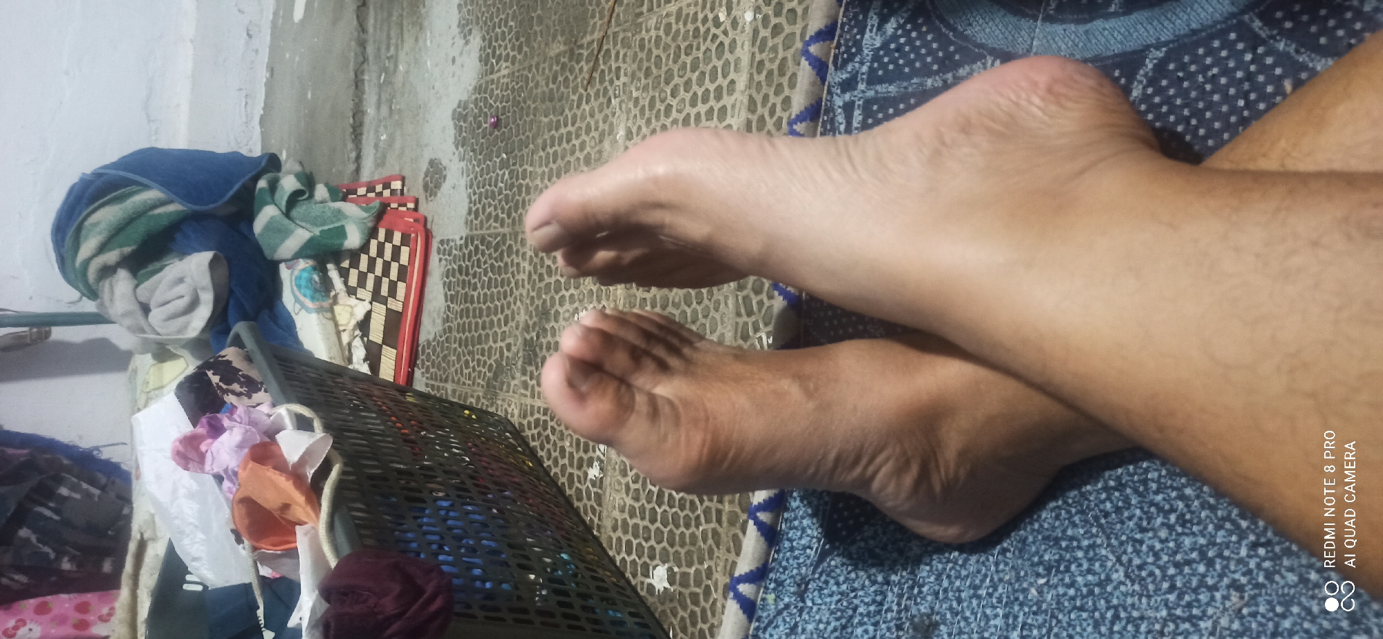 In Sedati Agung, Indonesia, a close-up shot focuses on a person's bare feet resting on a blue patterned cloth. The feet are crossed, with the soles visible, showing skin texture and toes. In the background, a laundry basket filled with various clothing items is partially visible. Beyond the basket, a tiled floor and a stack of folded towels and blankets are present, suggesting a domestic setting, possibly a laundry area. The lighting is dim, creating shadows and highlighting the texture of the floor and skin.
