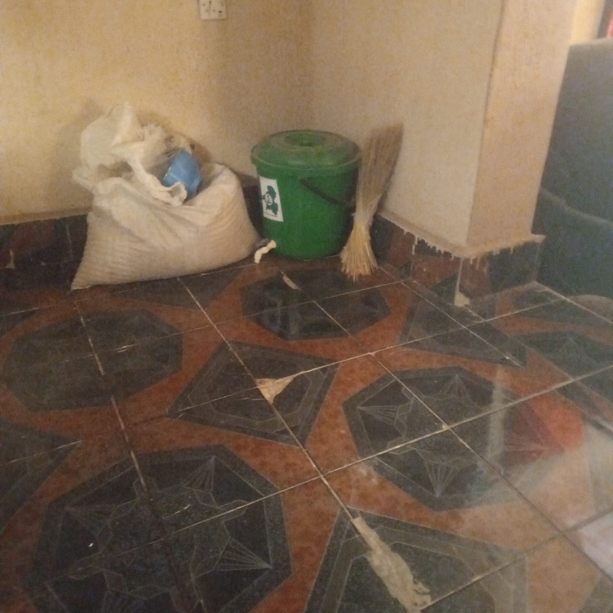 The image shows a corner of a room with tiled flooring. In the foreground, the floor is covered with dark and reddish-brown hexagonal tiles with a geometric pattern. In the background, against a light yellow wall, are several objects. On the left is a large white bag, possibly containing grain or sand, with a blue object partially visible inside. Next to the bag is a green plastic bucket with a lid, a white tap protruding from its side, and a sticker with a black and white graphic on its front. To the right of the bucket is a broom with straw bristles leaning against the wall. The wall appears to be textured and painted a light yellow color. There is a white electrical outlet visible on the upper left portion of the wall. The scene appears to be indoors, likely in a domestic or storage space. There are no people visible in the image. The overall impression is one of a utilitarian or storage area within a building. It's not possible to determine the exact location within Zinna, Nigeria from the image alone.