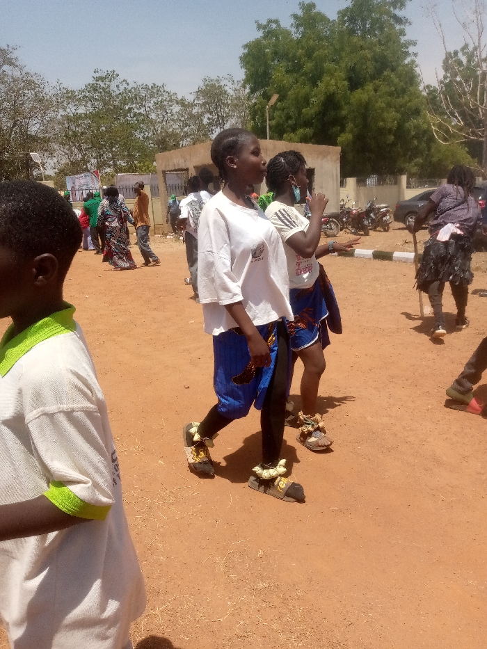 The scene depicts an outdoor gathering or procession in Kagoma, Nigeria, under clear, bright daylight. The ground consists of reddish-brown dirt or sand.
In the mid-foreground, two young females are walking from left to right. The leading female wears a white t-shirt with a dark logo, blue patterned shorts over black leggings, and black sandals adorned with multi-colored, possibly beaded, ankle wraps. She carries a small, dark, curved object in her right hand. The following female wears a striped white t-shirt, blue patterned shorts over black leggings, and sandals with similar multi-colored ankle adornments. She looks towards the right.
To the immediate left foreground, the partial profile of a young person in a white t-shirt with a lime green collar and sleeve trim is visible, facing right.
In the mid-ground and background, numerous individuals are present, walking or standing. One person on the far right wears dark clothing, a dark skirt-like garment, and holds a long stick, also displaying similar ankle adornments.
Architectural elements include a light-colored, single-story building with a flat roof. Several motorcycles are parked near a curb, behind which a dark sedan is also visible. Utility poles and a solar panel are present. Trees with varying foliage line the background. Posters or banners are partially visible on structures in the far-left background. Shadows are pronounced, indicating direct sunlight.