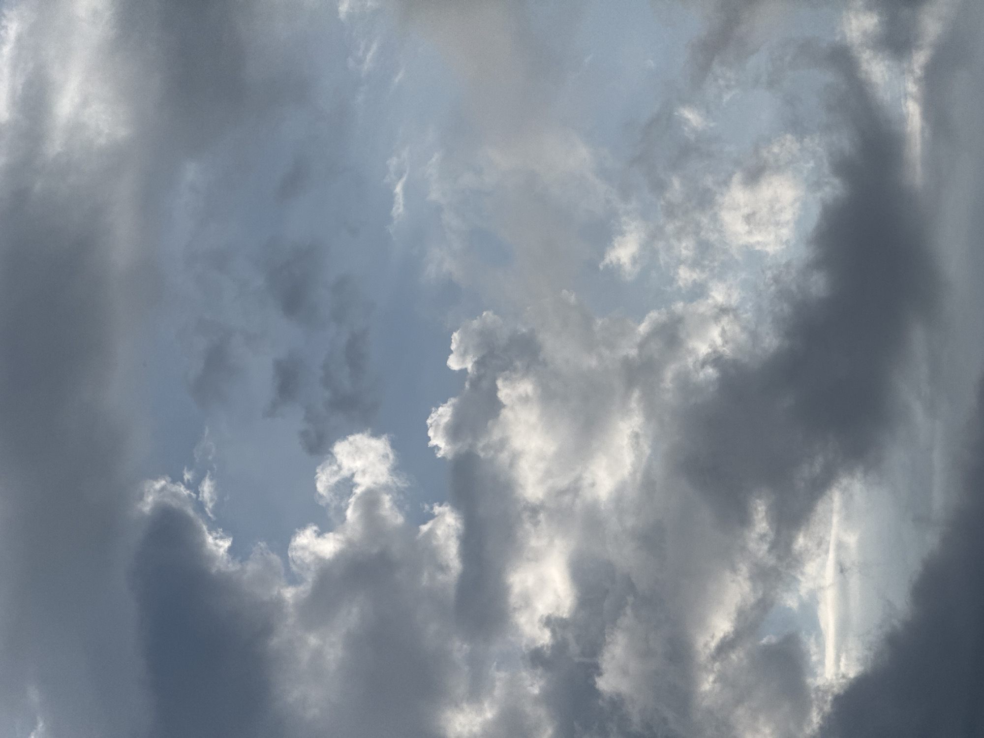 The image shows a cloudy sky above the city of Sary-Oy, Kyrgyzstan. The clouds are a mix of dark gray and white, with patches of blue sky visible in between. The sunlight is breaking through some of the clouds, creating bright highlights. There are no people or objects visible in the image, only the sky.