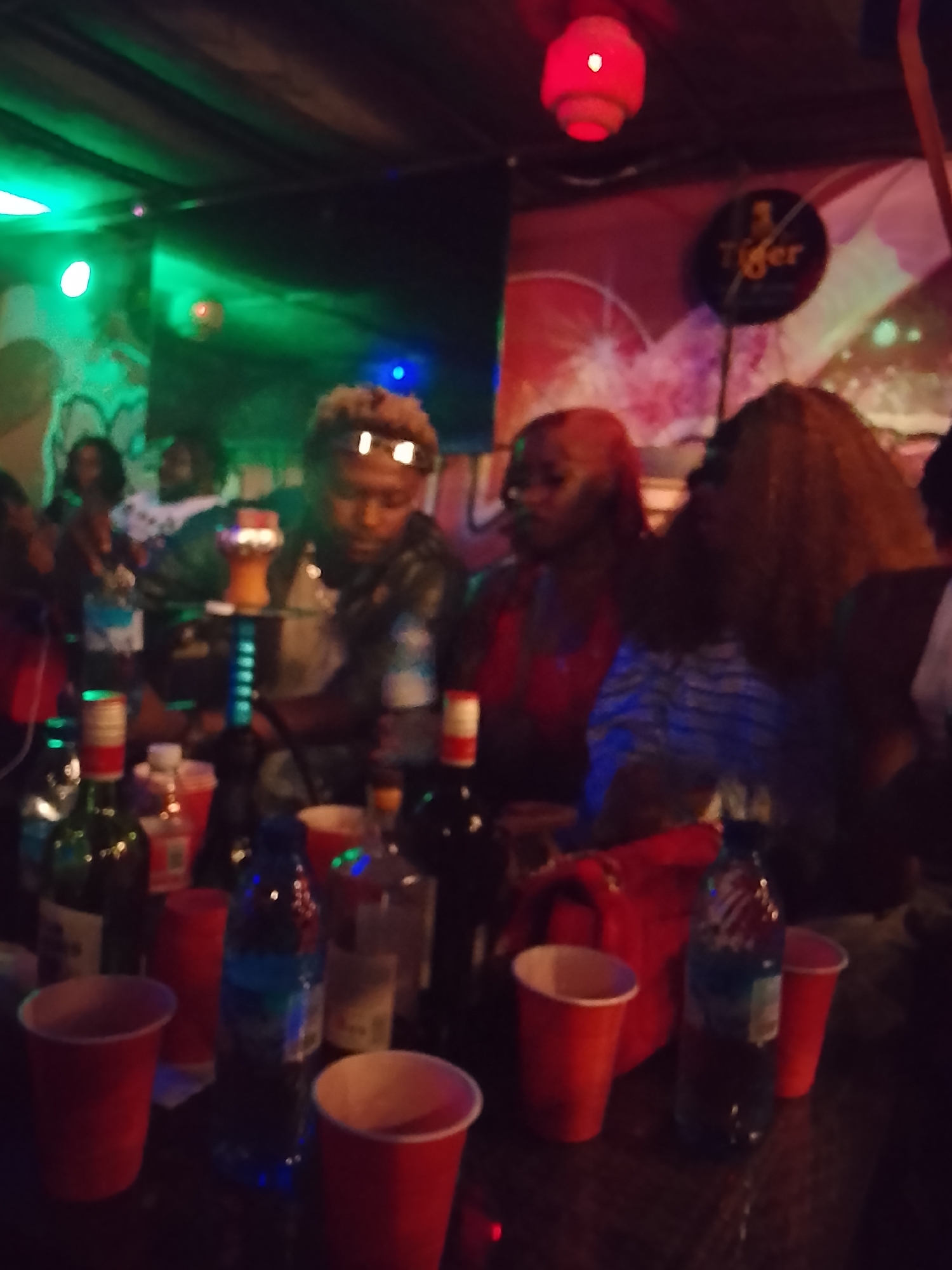 The image appears to be taken indoors in what might be a bar or club setting in Narayi, Nigeria. Several people are present, including a man with light-colored hair wearing sunglasses and at least two women. A hookah pipe is visible on the table in front of the man. There are numerous bottles and red plastic cups scattered on the table. The lighting is low and colorful, with green, blue, and red hues. A "Tiger" beer sign is visible on the wall.