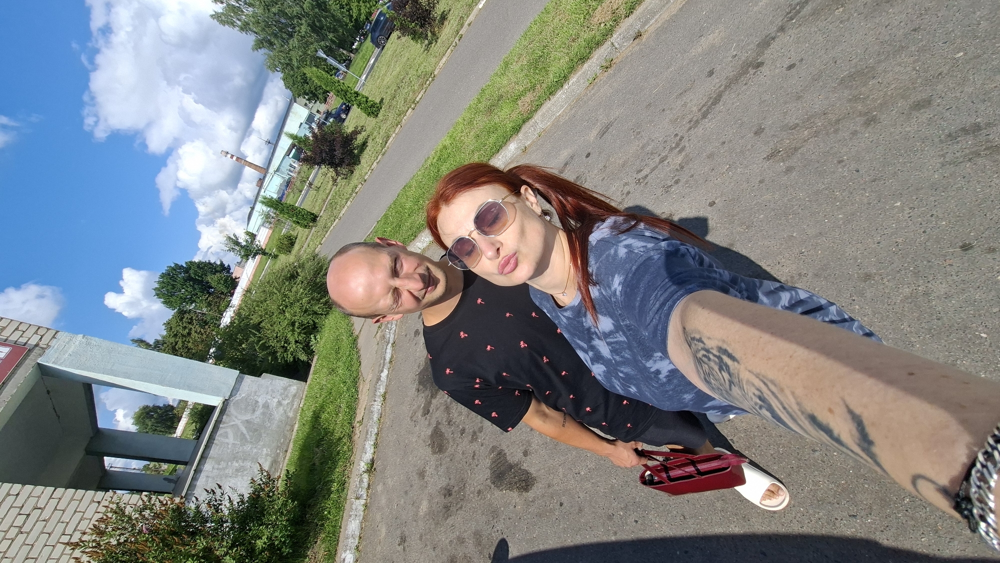 A couple, a bald man and a woman with red hair, are taking a selfie outdoors in the city of Myadzyel, Belarus. The man is wearing a black t-shirt with a palm tree pattern and is winking at the camera. The woman is wearing sunglasses and a blue tie-dye t-shirt, and she is puckering her lips. She is holding a red handbag. The background shows a building with a blue pillar, green bushes, and a cloudy blue sky. There is also a road and some trees visible. The image appears to be taken on a sunny day.