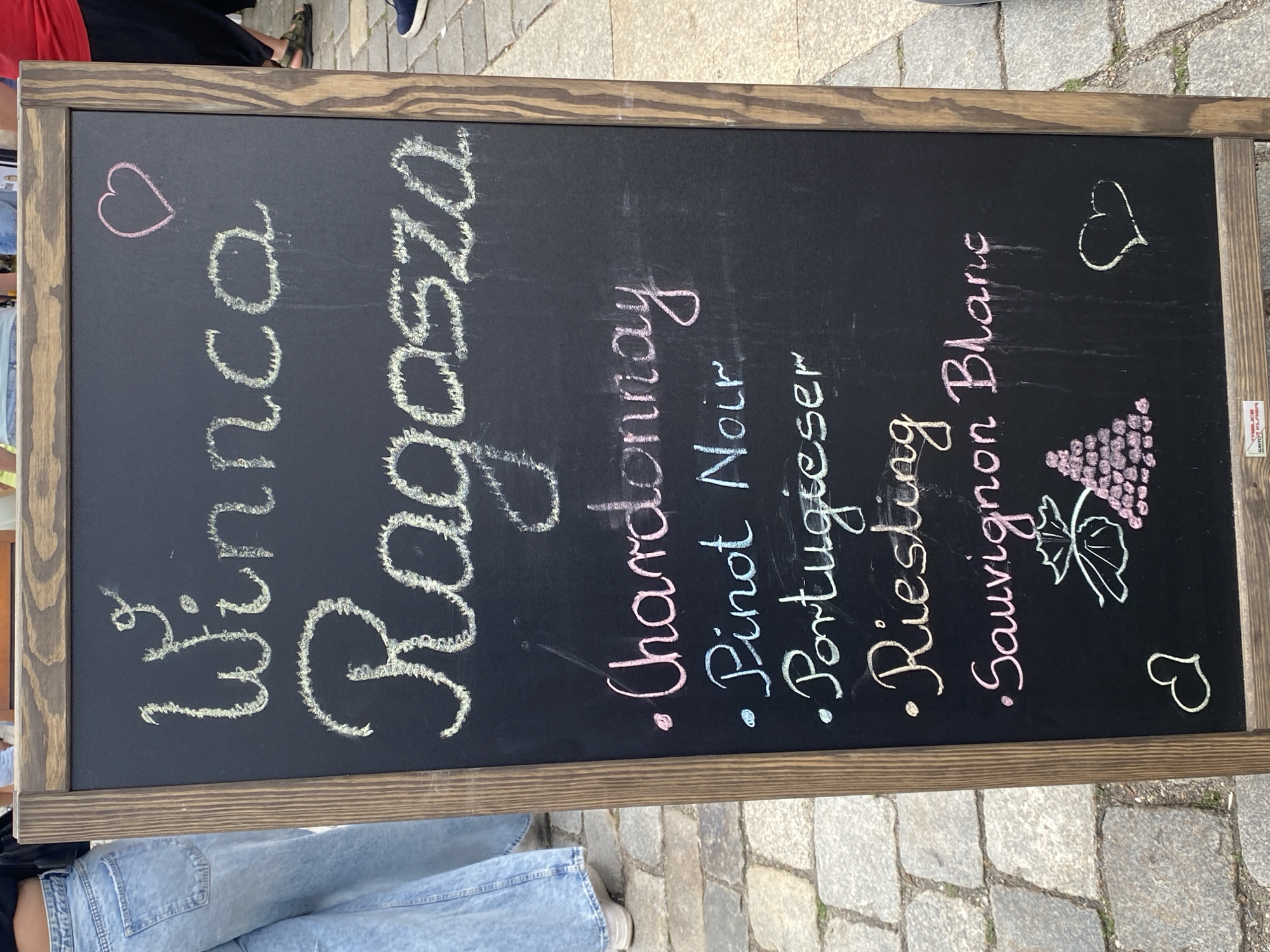 The image displays a freestanding chalkboard sign, positioned outdoors on a paved surface, likely in a pedestrian area in Wrocław, Poland. The sign has a dark wooden frame and a black chalk surface.
The top of the sign features the handwritten text "Winnica Ragosza" in white chalk. A small pink chalk heart is drawn in the top right corner of the board. Below the winery name, a list of five wine types is presented with bullet points:
* "Chardonnay" is written in pink chalk.
* "Pinot Noir" is written in light blue chalk.
* "Portugieser" is written in light green chalk.
* "Riesling" is written in yellow chalk.
* "Sauvignon Blanc" is written in pink chalk.
At the bottom of the sign, on the left, a chalk drawing depicts a green vine leaf with a cluster of pink grapes. On the bottom right, a white chalk outline of a heart is drawn. The chalk writing exhibits some smudging and faint vertical drip marks.
The ground beneath and around the sign consists of light grey cobblestones on the left side and a smoother, darker grey paving on the right side.
On the left side of the frame, the lower body of a person is visible, wearing light blue, wide-leg denim jeans and white athletic shoes. On the right side, the lower legs and feet of another person are visible, wearing black trousers and dark, open-toed sandals (e.g., Birkenstock-style), with a hint of a red garment above. The background is slightly out of focus, showing indistinct shapes of other people and urban elements. The presence of the "Winnica Ragosza" name, which is a Polish winery, indicates the location is a point of sale for their products within Wrocław, Poland.