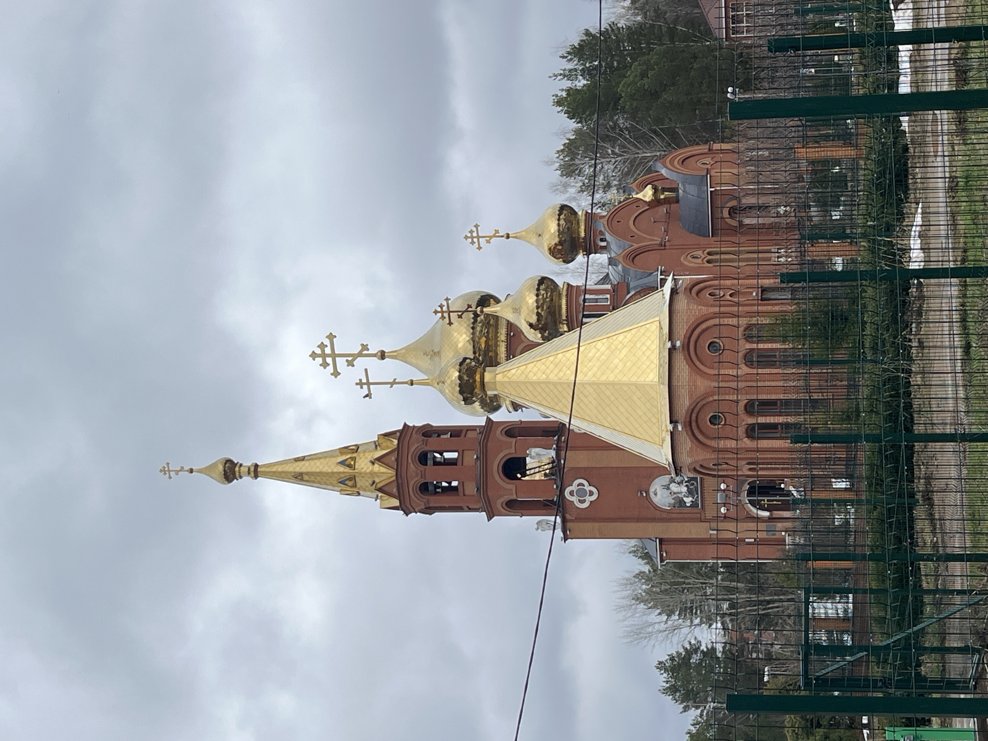 The image shows the Orthodox Church of St. George the Victorious in Chaikovsky, Russia. The church is constructed of red brick with gold domes and crosses. A green metal fence is in the foreground. The sky is overcast. Trees are visible in the background.