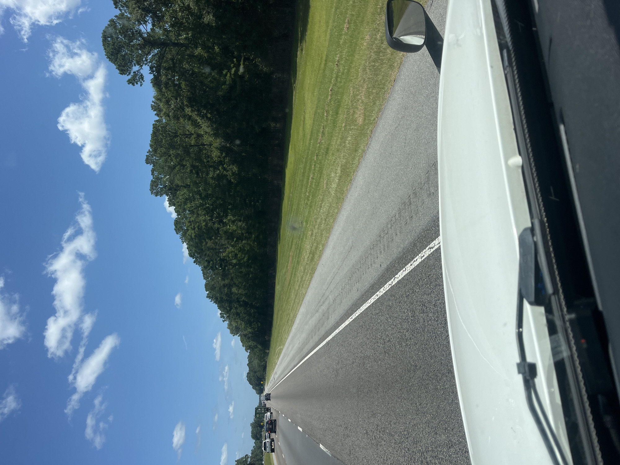 A white truck's dashboard is visible in the foreground, with windshield wipers and a portion of the hood. The view extends to a highway with several lanes and other vehicles, including cars and a truck. The highway is bordered by a grassy shoulder and a dense line of green trees. The sky is blue with scattered white clouds. The scene appears to be a daytime drive on a highway in or near Riceboro, United States.
