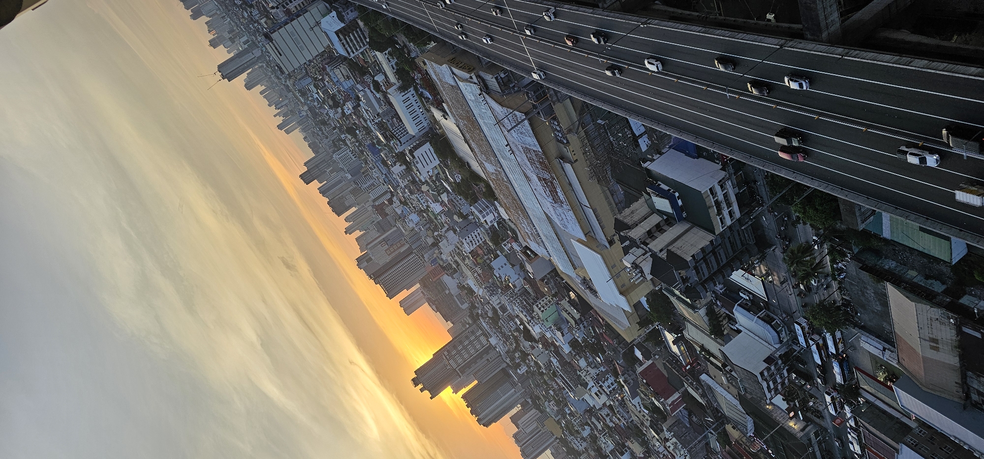 This is a high-angle, wide shot of a city skyline in Makati, Philippines, during a sunset. The sky is filled with hues of orange and yellow, with streaks of clouds extending across the frame. In the background, numerous skyscrapers of varying heights dominate the cityscape. A multi-lane elevated highway, with several cars and trucks traveling on it, cuts diagonally across the right side of the image. Below the highway and interspersed among the buildings are residential and commercial structures, some with visible rooftops and windows. The scene depicts a bustling urban environment under a twilight sky.