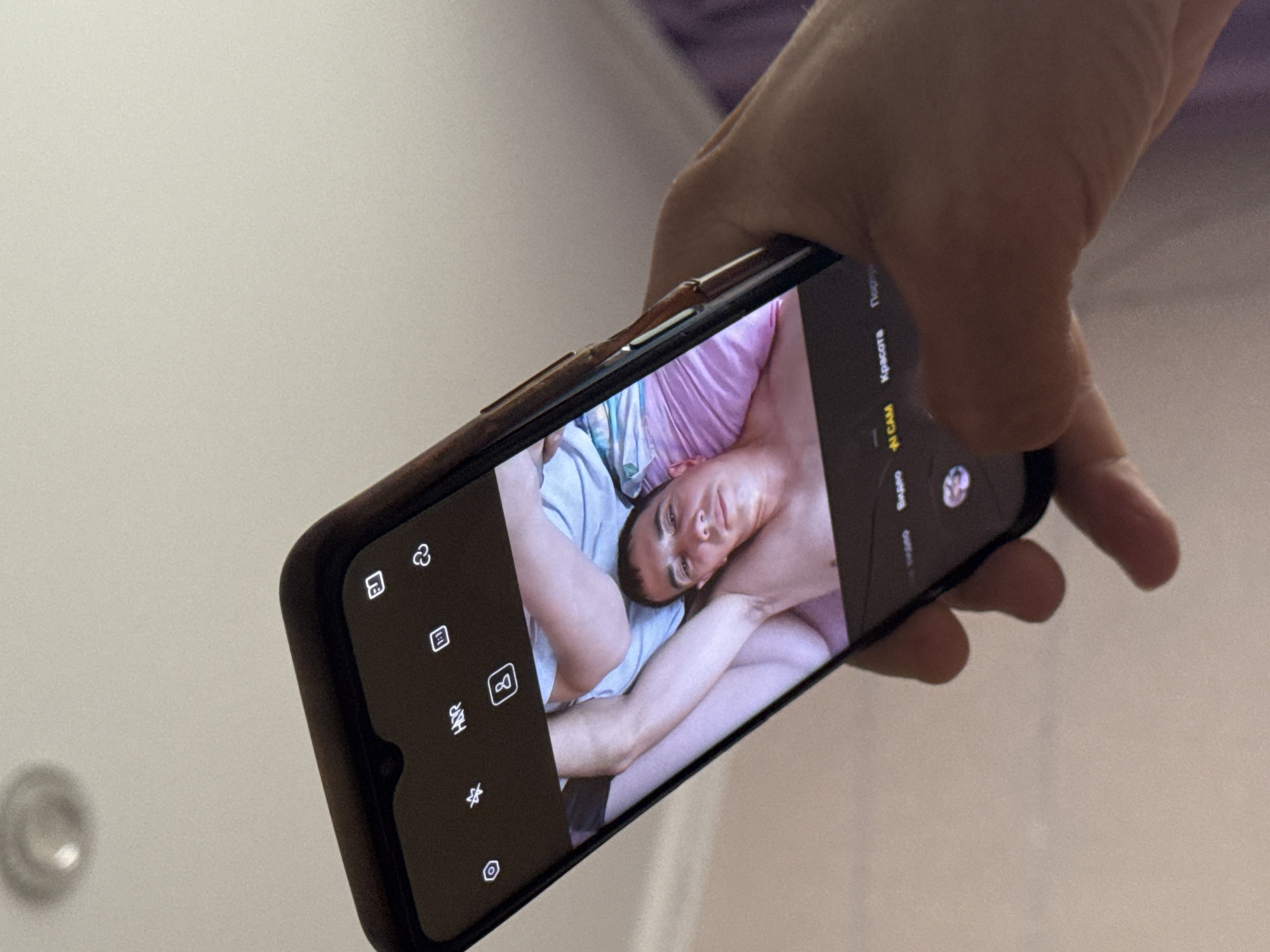 A hand holds a smartphone, displaying a selfie of two individuals. The person in the foreground of the selfie appears to be a man, lying down with his head resting on a pillow, his gaze directed towards the camera. Partially visible behind him is another person, whose legs are intertwined with his. The phone's screen also shows camera interface elements. The background is out of focus, suggesting an indoor setting. There is no information in the image to indicate the location in the city of Asha, Russia.