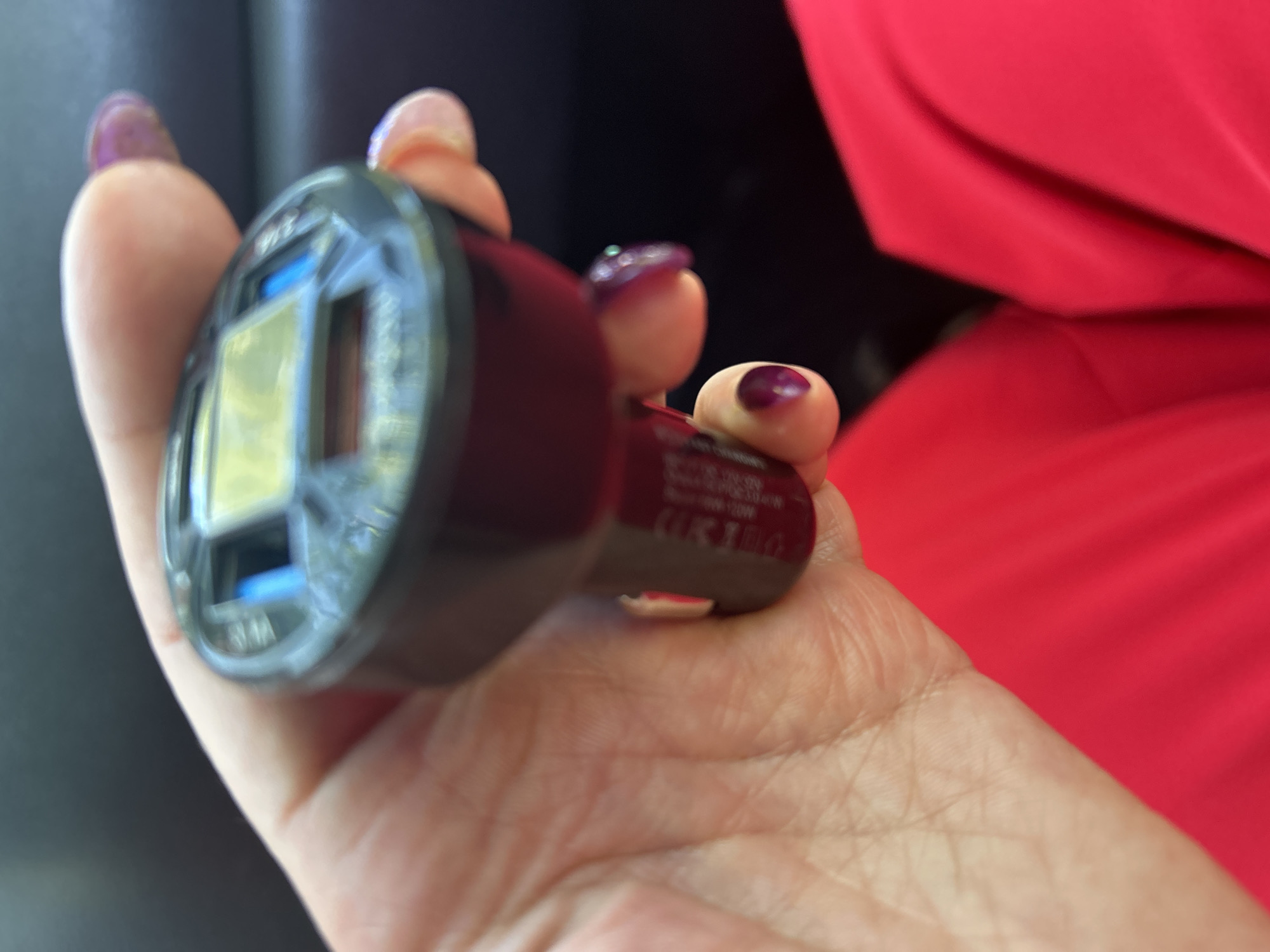 A close-up, dimly lit, angled shot shows a hand with purple nail polish holding a black car charger. The charger has a small, glowing yellow screen and two USB ports. The background is dark, suggesting an indoor or nighttime setting, possibly in a car. There is no discernible connection to Tugulym, Russia.