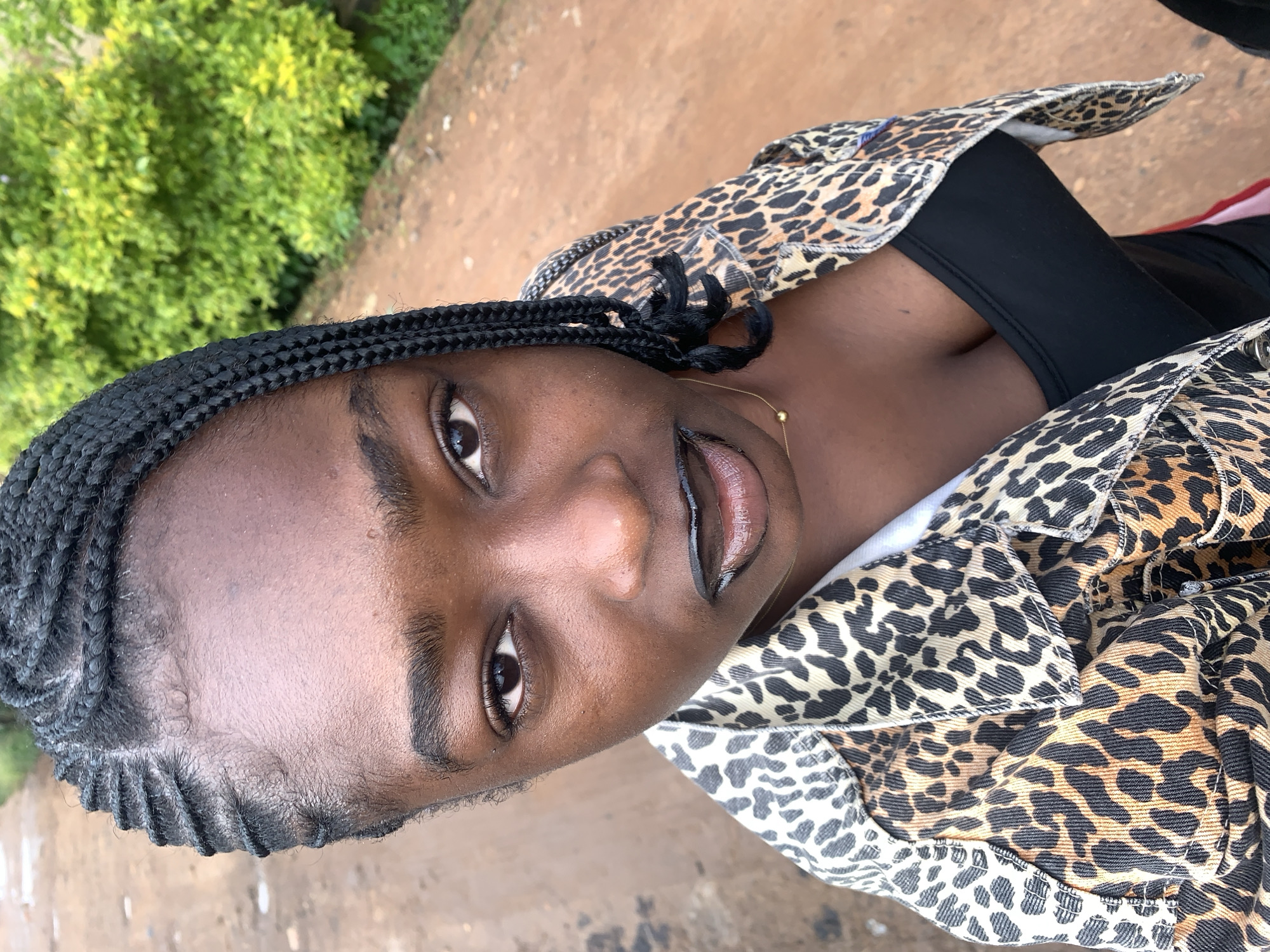 A close-up, high-angle outdoor portrait features a young Black woman with braided hair. She is wearing a black tank top and a leopard-print denim jacket. Her lips are painted with black lipstick. The background consists of a dirt path and lush green foliage. The scene appears to be located in Rafin Par, Nigeria.