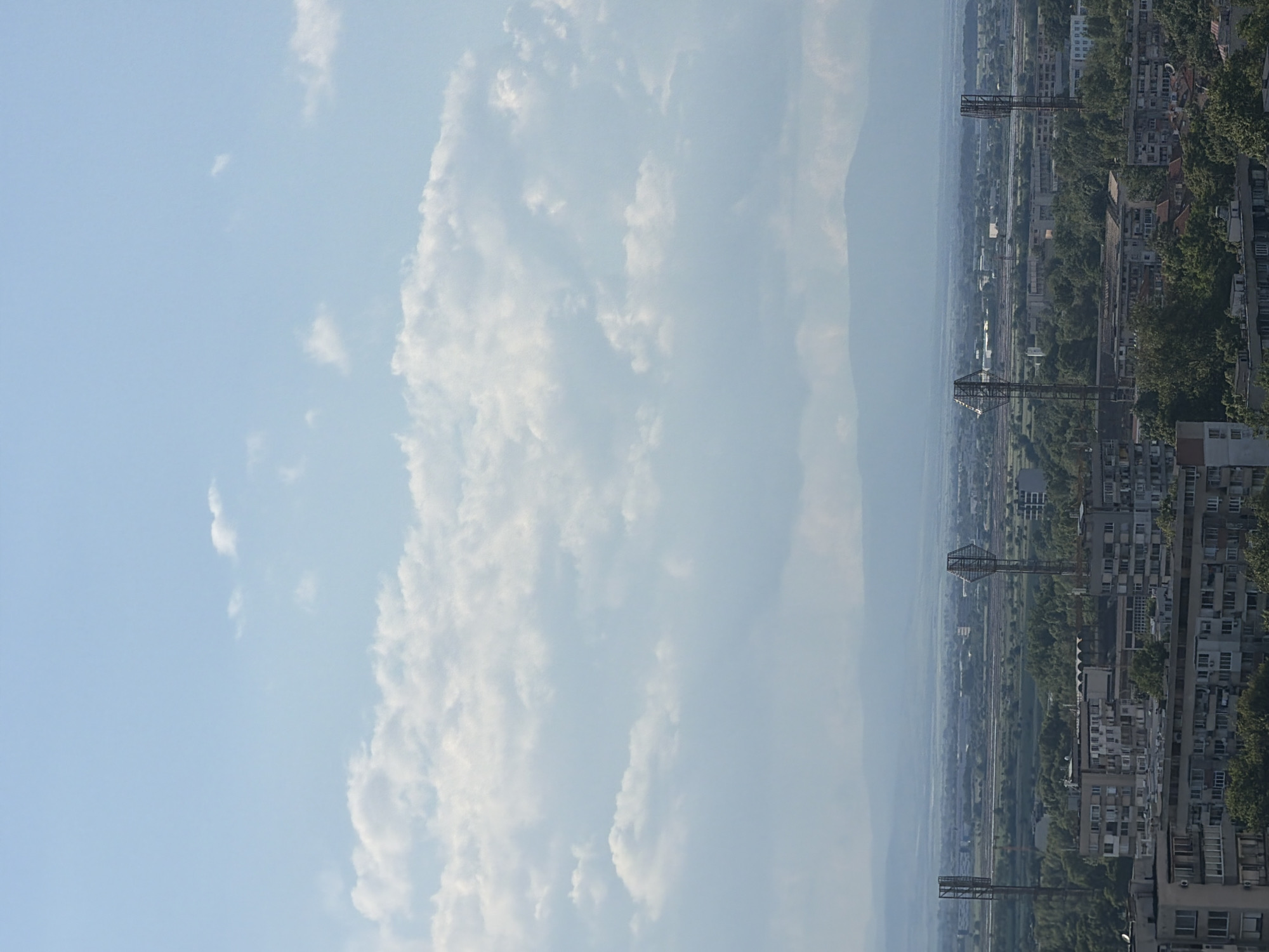 The image shows an elevated view of a cityscape in Plovdiv, Bulgaria. The right side of the image displays densely packed apartment buildings intermixed with greenery. Several metal utility towers and power lines run parallel to the buildings, indicating a railway or industrial area in the middle ground. The background appears hazy, obscuring details of distant mountains or hills. To the left, the sky is blue with substantial white clouds.