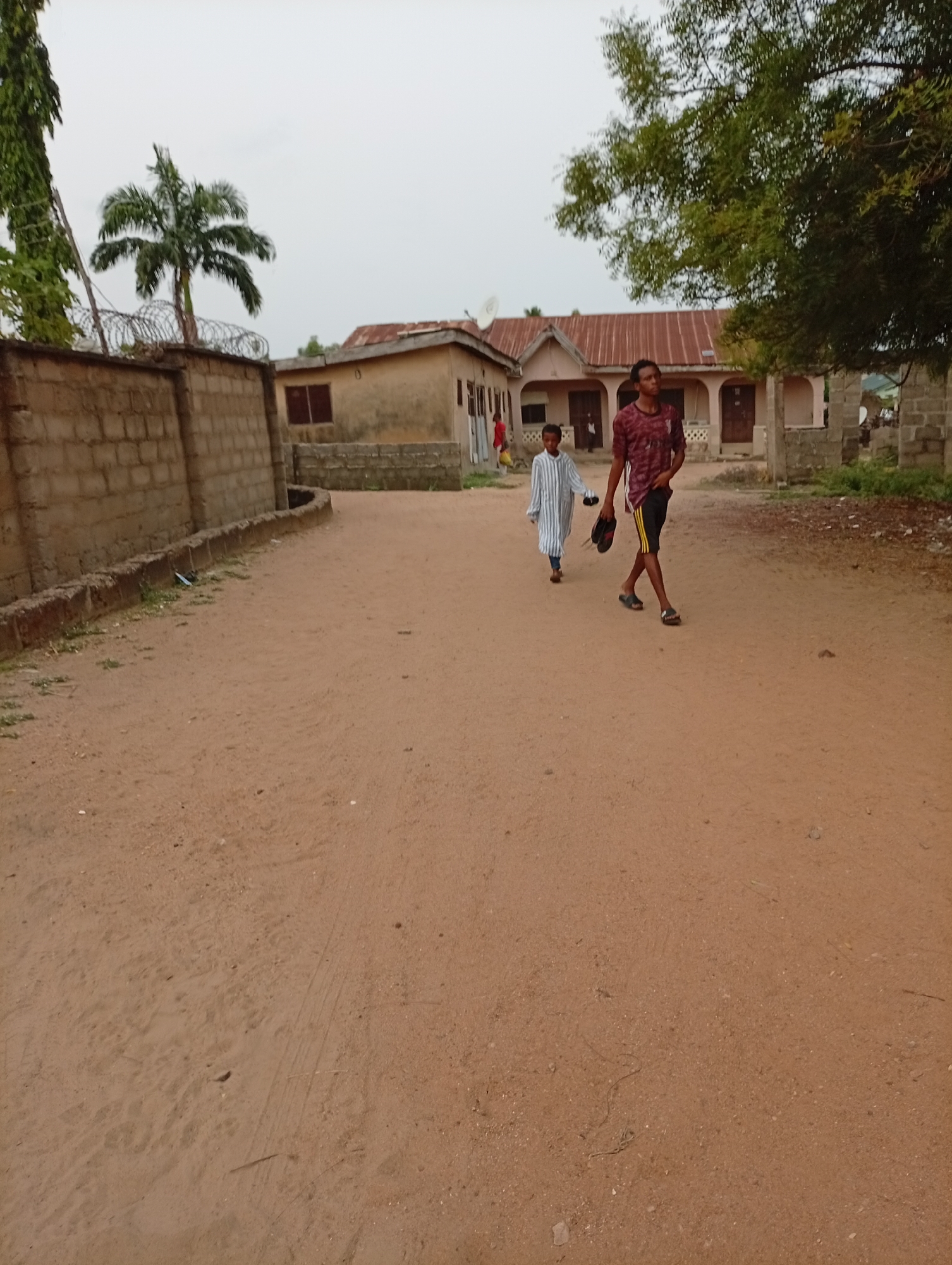 Location: Adankolo, Nigeria.
Two people are walking down a dirt road. The person in the front is wearing a maroon jersey and black shorts. They are holding a pair of shoes in their left hand. The second person is wearing a long-sleeved striped shirt. A wall with barbed wire on top is to the left of the road. Behind the two people are buildings with metal roofs and open doorways. There is also a tree to the right. The sky is overcast.