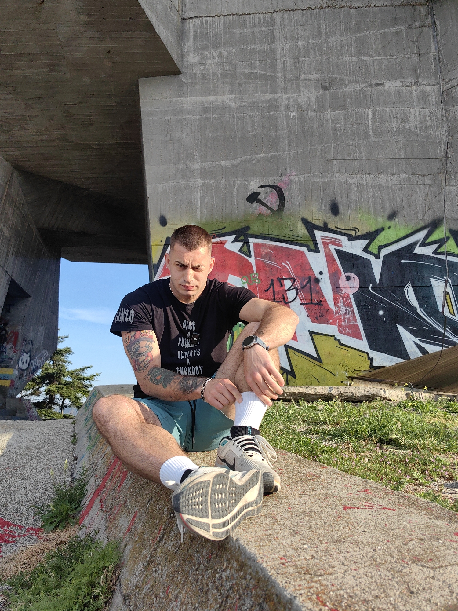 The image shows a man sitting on a concrete structure outdoors. The man has short hair, a t-shirt with graphic text, shorts, socks, and sneakers. He is wearing a watch on his left wrist and has tattoos on his left arm. Behind him is a large concrete wall with graffiti art. There are some plants growing around the concrete structure. The setting appears to be beneath a concrete overpass or bridge. The location is in the city of Unknown, Unknown.