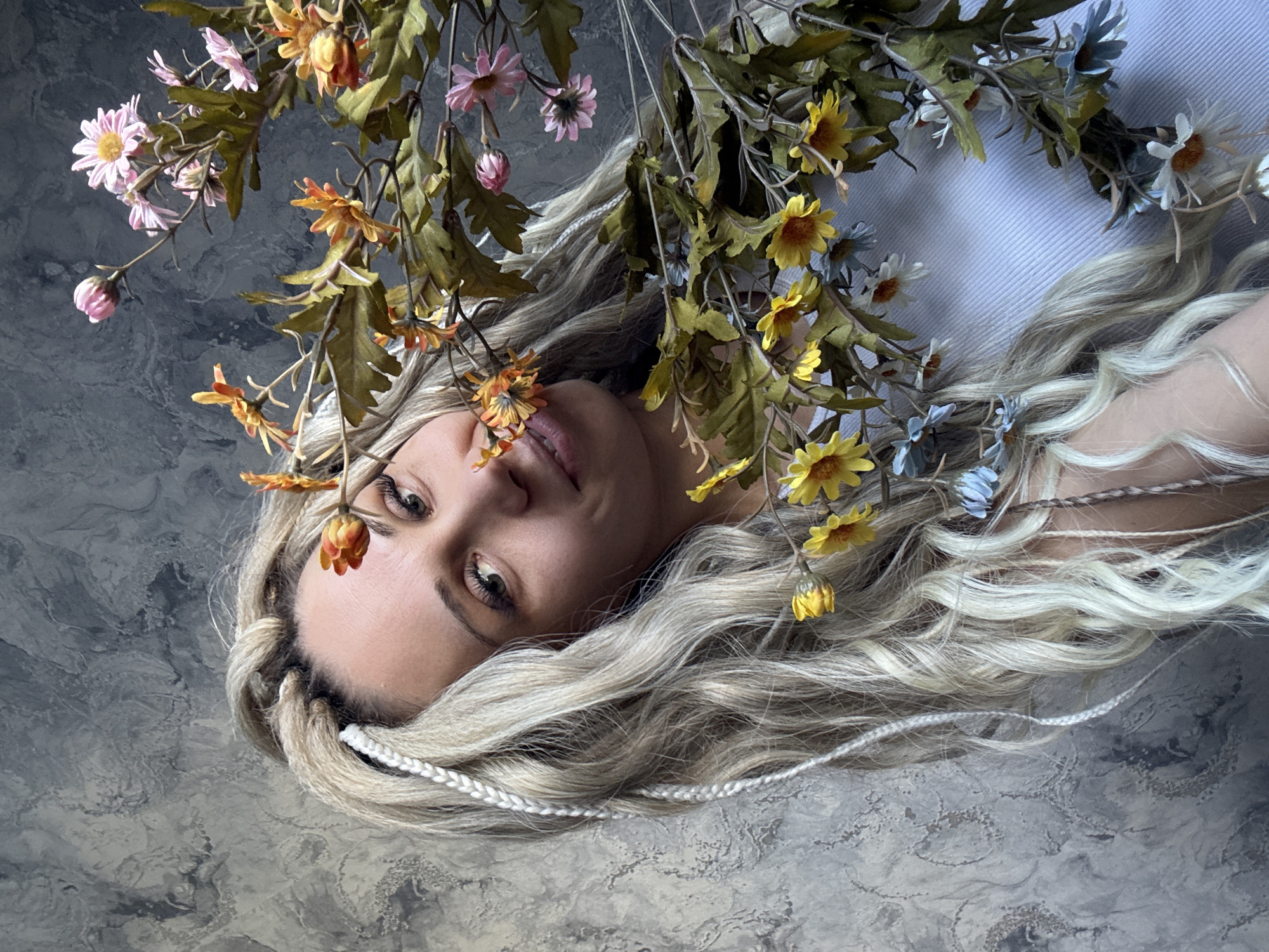 A blonde woman lies on a grey mottled surface, her head positioned in the top left corner of the frame. She is adorned with a floral arrangement featuring orange, yellow, pink, and blue flowers. Her hair is styled with braids. The background is a mottled grey, possibly a textured surface. The scene is not explicitly identifiable as Rusanovo, Russia, based on the visual information provided.