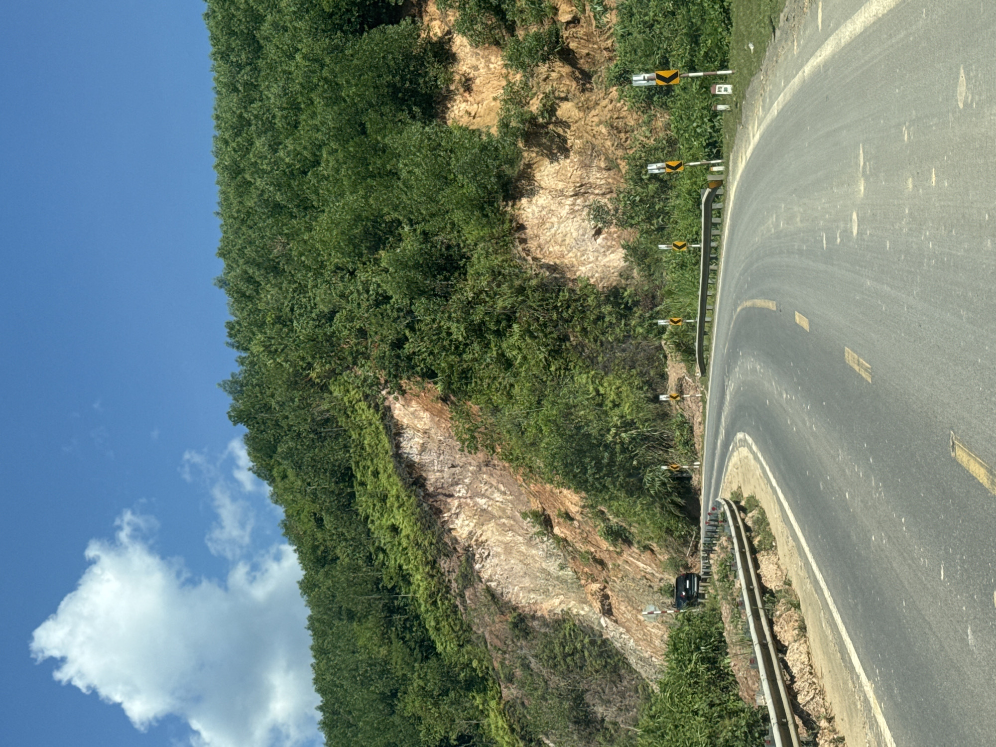 The image depicts a winding asphalt road curving to the right, with a steep, vegetated hillside on the left. The hillside features a prominent section of exposed, reddish-brown and light-colored rock face, indicating a recent landslide or erosion. A metal guardrail runs along the edge of the road, separating it from the slope. Several yellow and black directional warning signs are visible along the roadside, indicating curves and potential hazards. A black SUV is parked on the shoulder of the road on the left side, near the guardrail. The sky is bright blue with a few scattered white clouds. The environment appears to be a rural or mountainous area. This scene is consistent with the topography and road infrastructure found in mountainous regions of Vietnam, such as the province of Khánh Hòa, where Nam Khánh Vĩnh is located.
