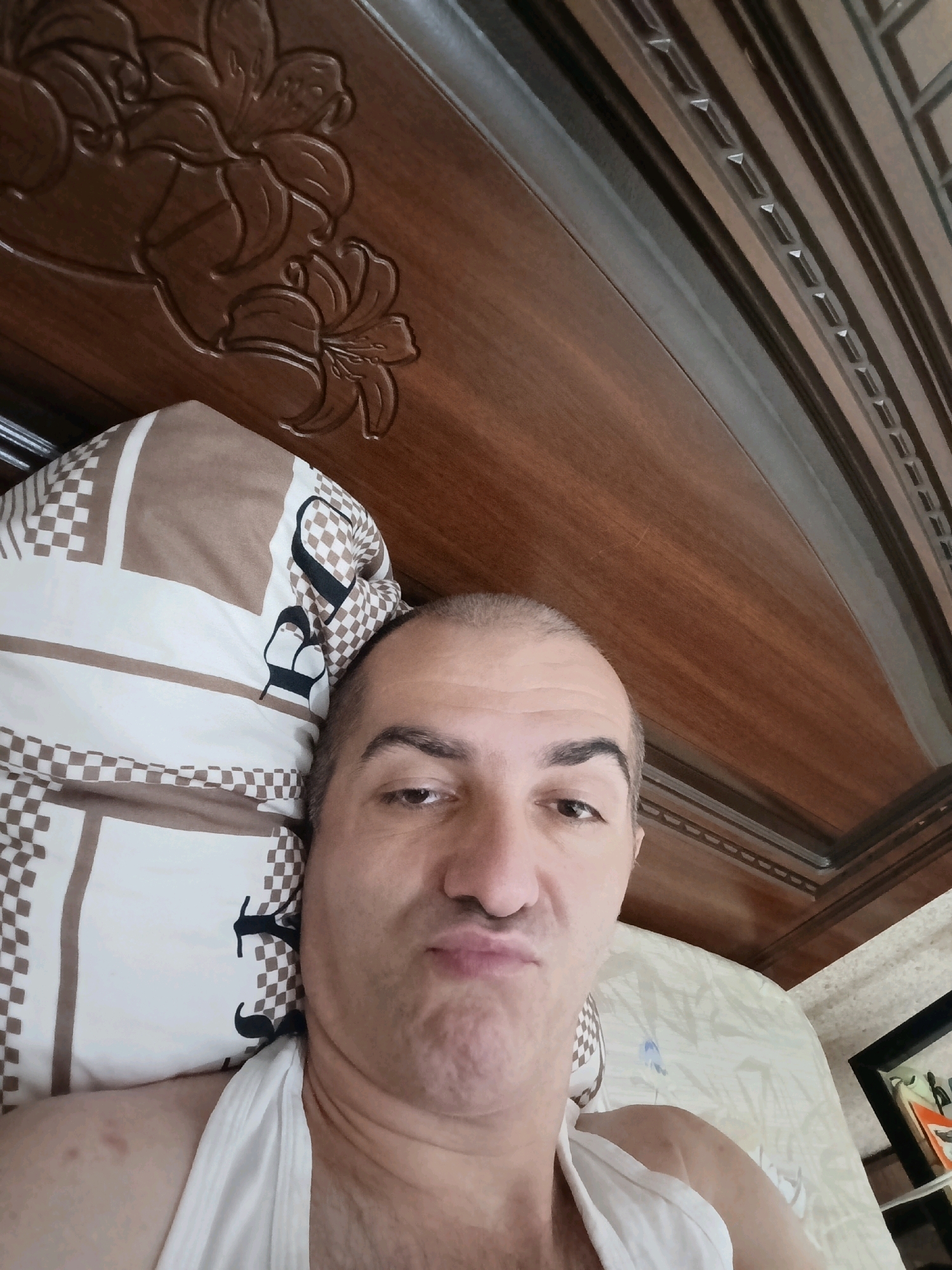 A man is lying on a bed, taking a selfie. He is wearing a white tank top. The bed has a wooden headboard with carved floral designs. A pillow with a checkered and patterned design is visible. The scene appears to be indoors, possibly in a bedroom. There is no indication of the location being Grozny, Russia.