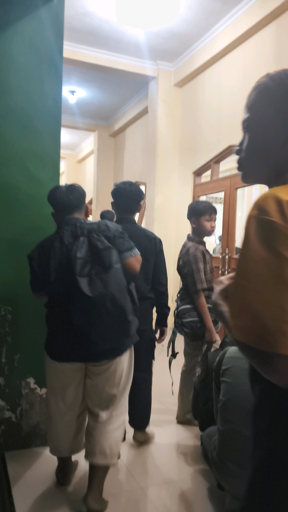 The image captures an indoor scene, likely a hallway or corridor, with several individuals present. The lighting suggests an artificial, possibly fluorescent, source. The walls appear to be painted in light, neutral tones, with a green wall visible on the left. A wooden door with glass panels is situated in the background.
Several people are seen from behind or in profile. One individual in the foreground on the left wears a black shirt and light-colored pants and carries a black backpack. To their right, another person in a black shirt is visible. Further down the hallway, a younger person in a checkered shirt is standing, and another individual, partially obscured, is wearing a yellow top. The floor is tiled, and shadows are cast on it. The overall impression is of a group of people in transit or gathered in an indoor public or semi-public space. The context of Trucuk, Indonesia, suggests this could be a school, community center, or a similar facility.