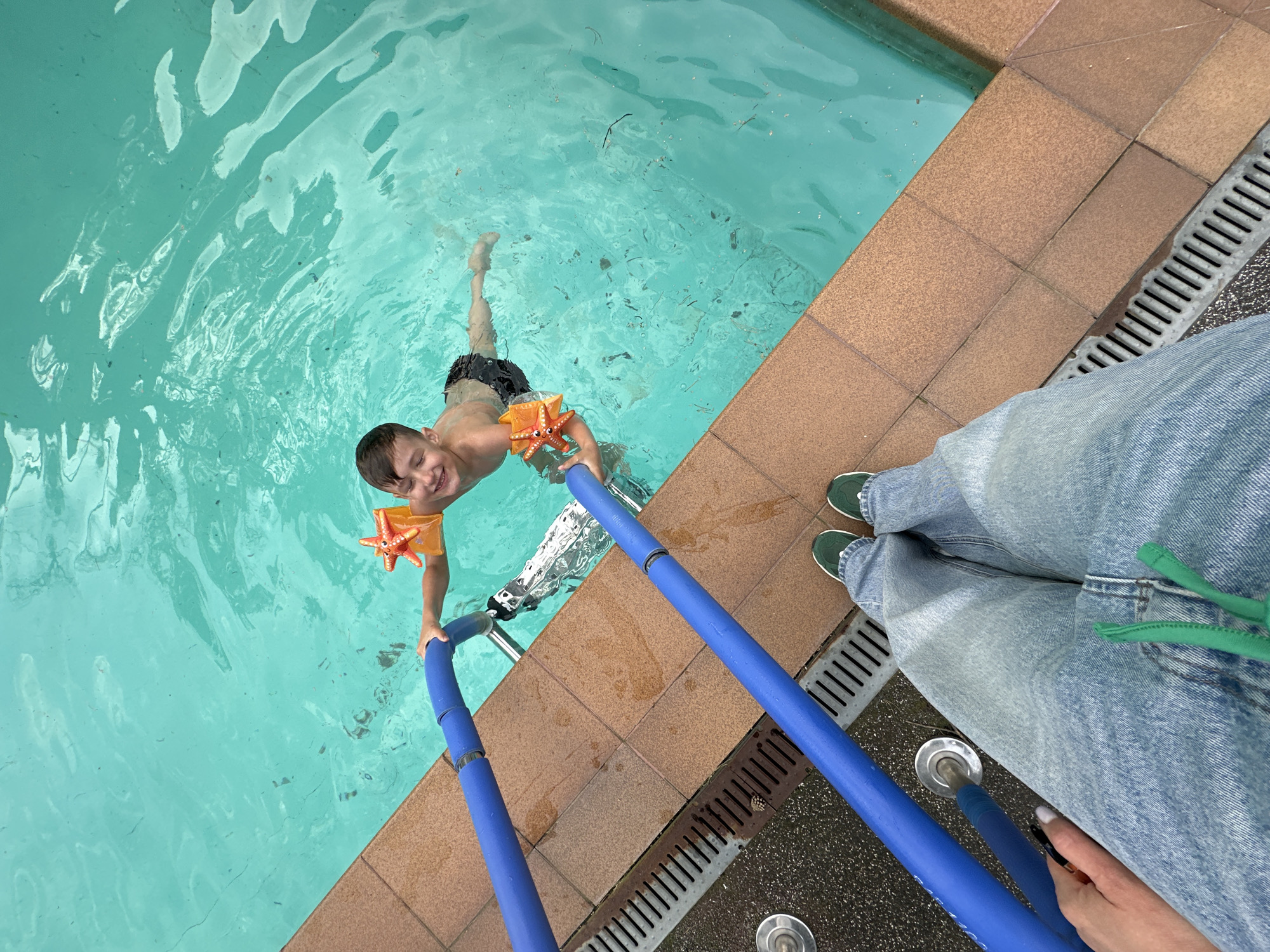 A child with brown hair, wearing black swim trunks and orange starfish-shaped arm floaties, is in a swimming pool. He is holding onto a blue pool noodle that is connected to a metal pool ladder. The water is turquoise. The pool has a tile border and a drainage grate. The lower legs and feet of a person wearing green shoes and light blue jeans are visible next to the pool. A hand is holding the end of the pool noodle. The location is not identifiable as Chaikovsky, Russia, from the provided visual information.