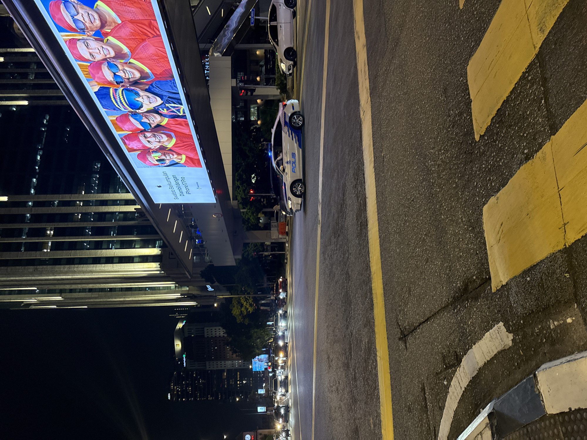 The image captures a street scene at night in Kuala Lumpur, Malaysia, featuring an illuminated advertising billboard showing images of swimmers in red swim caps. A white Malaysian police car ("POLIS") is parked on the street, and other cars are visible. The street has yellow and white painted lane markings. Tall buildings can be seen in the background, indicating an urban setting.