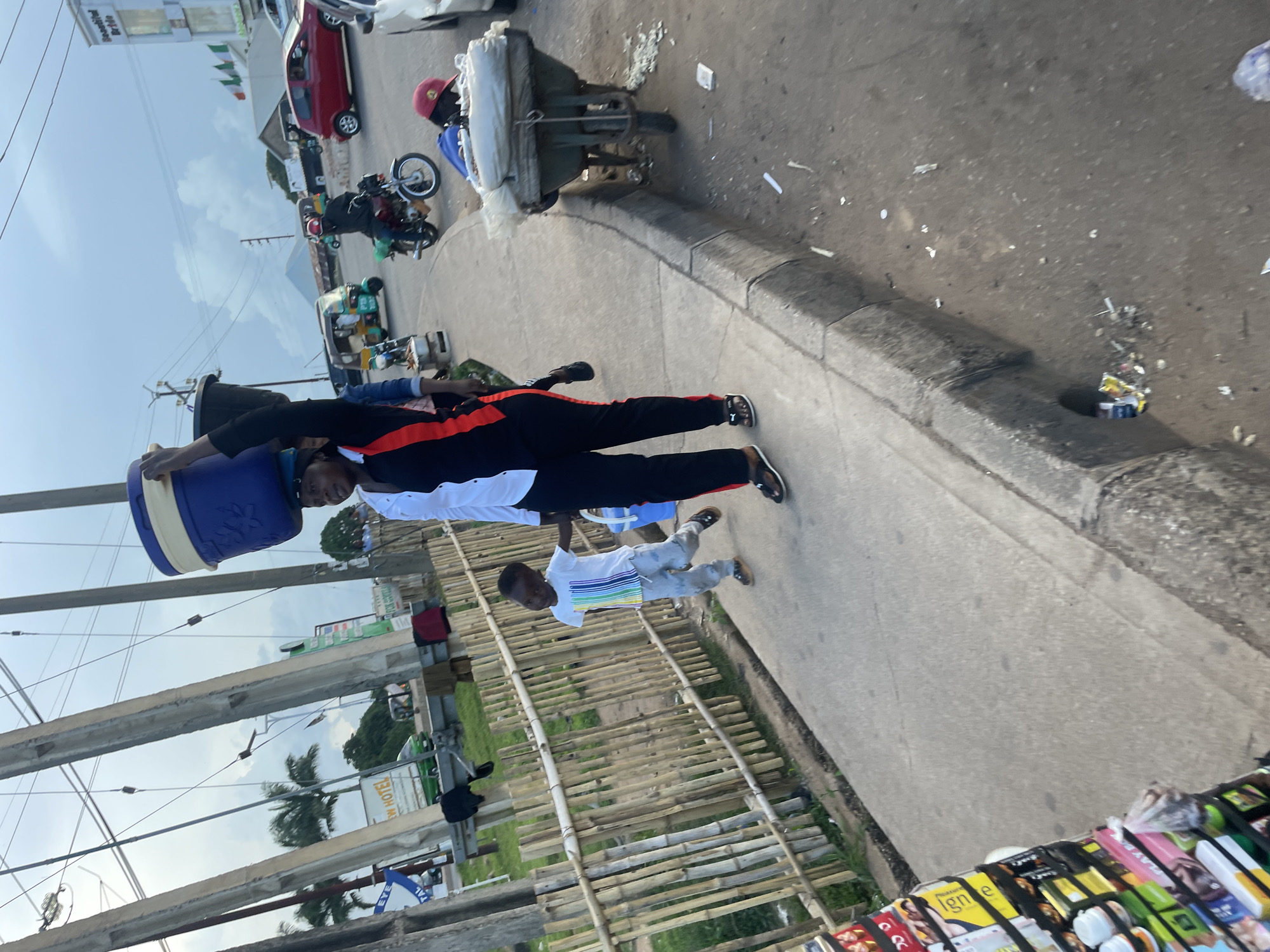The image depicts a street scene in Jos, Nigeria. A woman is walking, balancing a large blue and white cooler on her head. She is wearing a black and red outfit. Next to her, a young boy in a white t-shirt and grey pants walks along the sidewalk, holding her hand. In the background, there is a bamboo fence, a road with various vehicles including a red car, a motorcycle, and a three-wheeled auto-rickshaw. Power lines and poles are visible overhead, along with buildings and trees. To the right, a wheelbarrow is parked on the side of the road, loaded with materials wrapped in plastic. The ground is paved with concrete on the sidewalk and asphalt on the road, with some debris scattered on the ground. The sky is overcast.