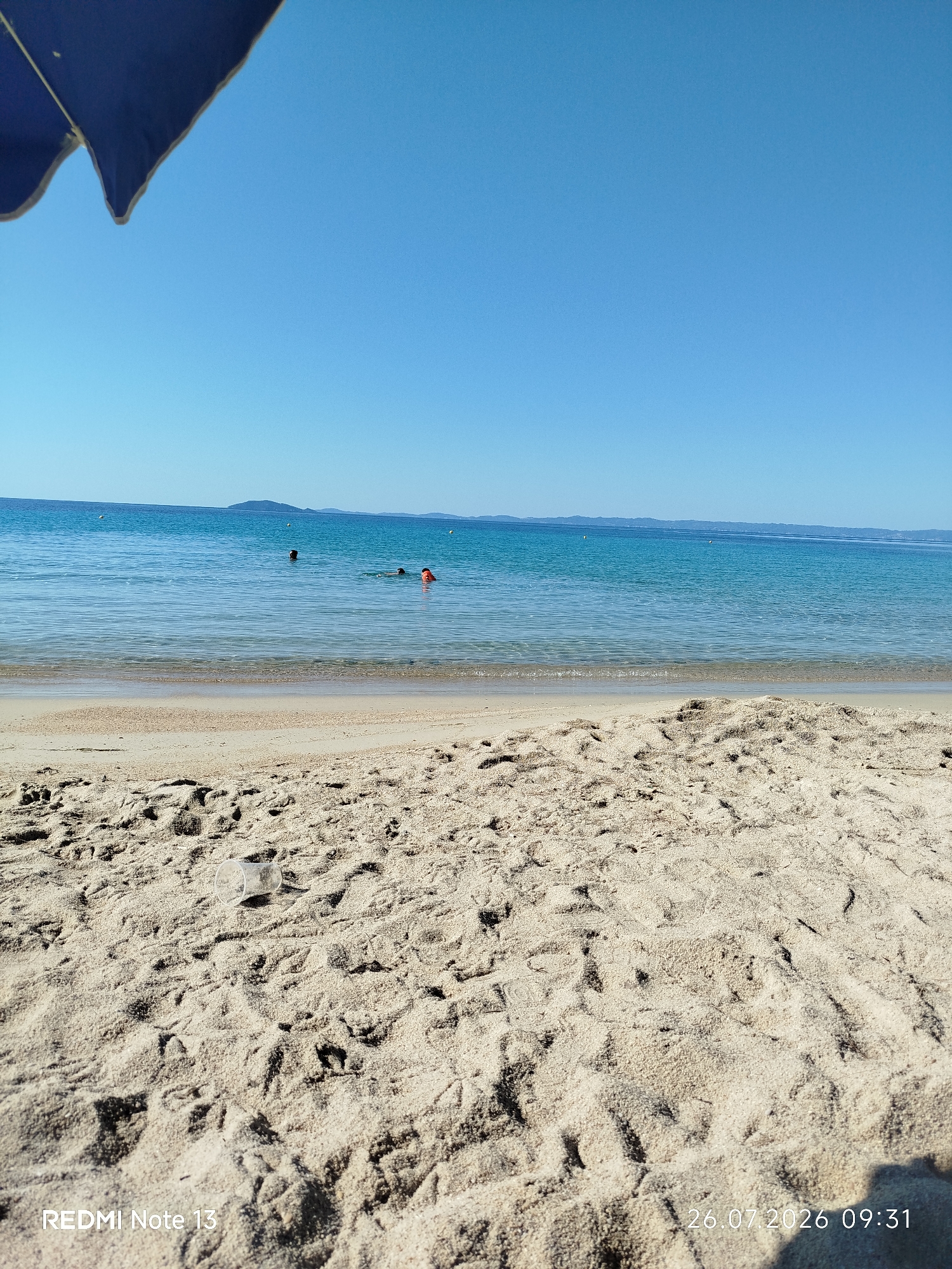 The image shows a sunny day at a sandy beach in Sithonia, Greece. In the foreground, there is a textured sandy beach with footprints and a clear plastic cup lying on the sand. A blue umbrella partially obscures the top left corner of the frame. The middle ground features the calm, clear turquoise water of the sea, with several people swimming. Two individuals are visible in the water, one with an orange buoy. In the background, a distant coastline with hills is visible under a clear blue sky. The date and time stamp "26.07.2026 09:31" and the text "REDMI Note 13" are visible in the lower part of the image.