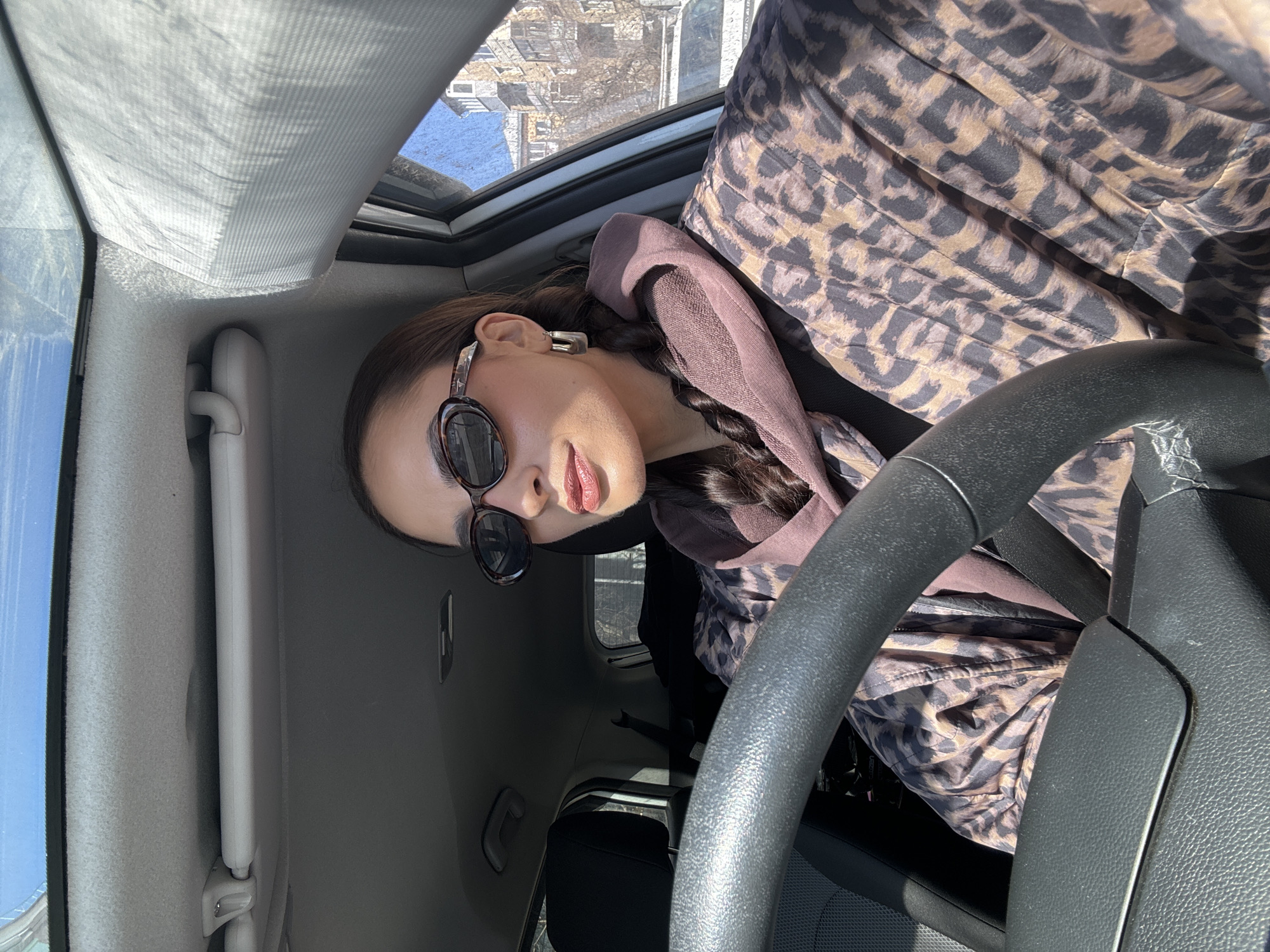 A woman sits in the driver's seat of a car. She is wearing sunglasses and a hooded jacket with a leopard print. The car has a sunroof. Outside the sunroof, buildings can be seen, likely in Krasnoyarsk, Russia. The woman's face is partially visible. Her hair is in a braid. The steering wheel is prominently in the foreground. The car's interior is light gray.