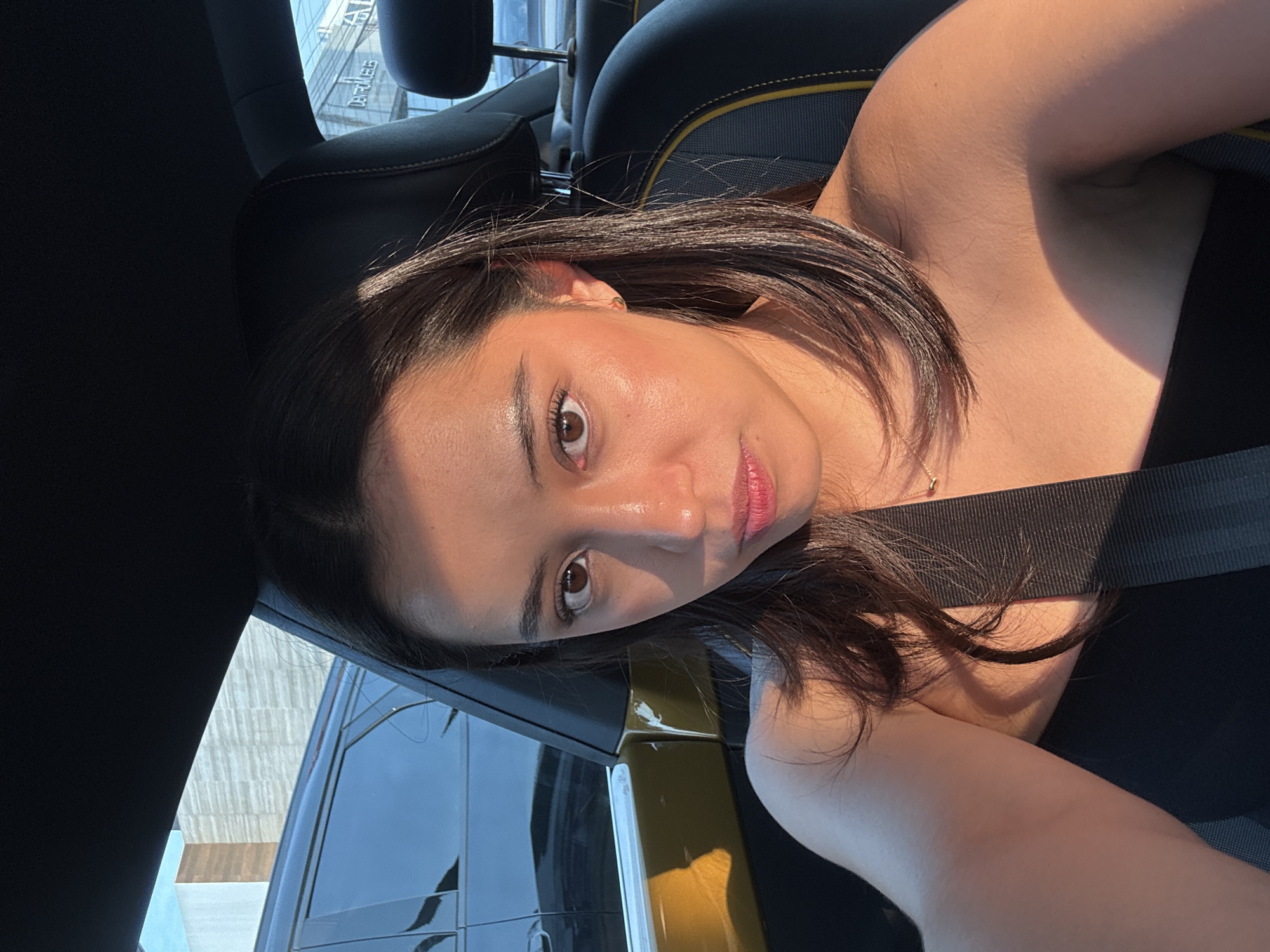 A woman is seated in a car, wearing a seatbelt. She has short, dark hair, brown eyes, and light skin. She is facing the camera with a slight angle. The car's interior is black with a yellow accent on the seat. Through the car window, a modern building with "CENTRO PUEBLA" visible is partially seen. This indicates that the location is likely in the city of Puebla, Mexico.