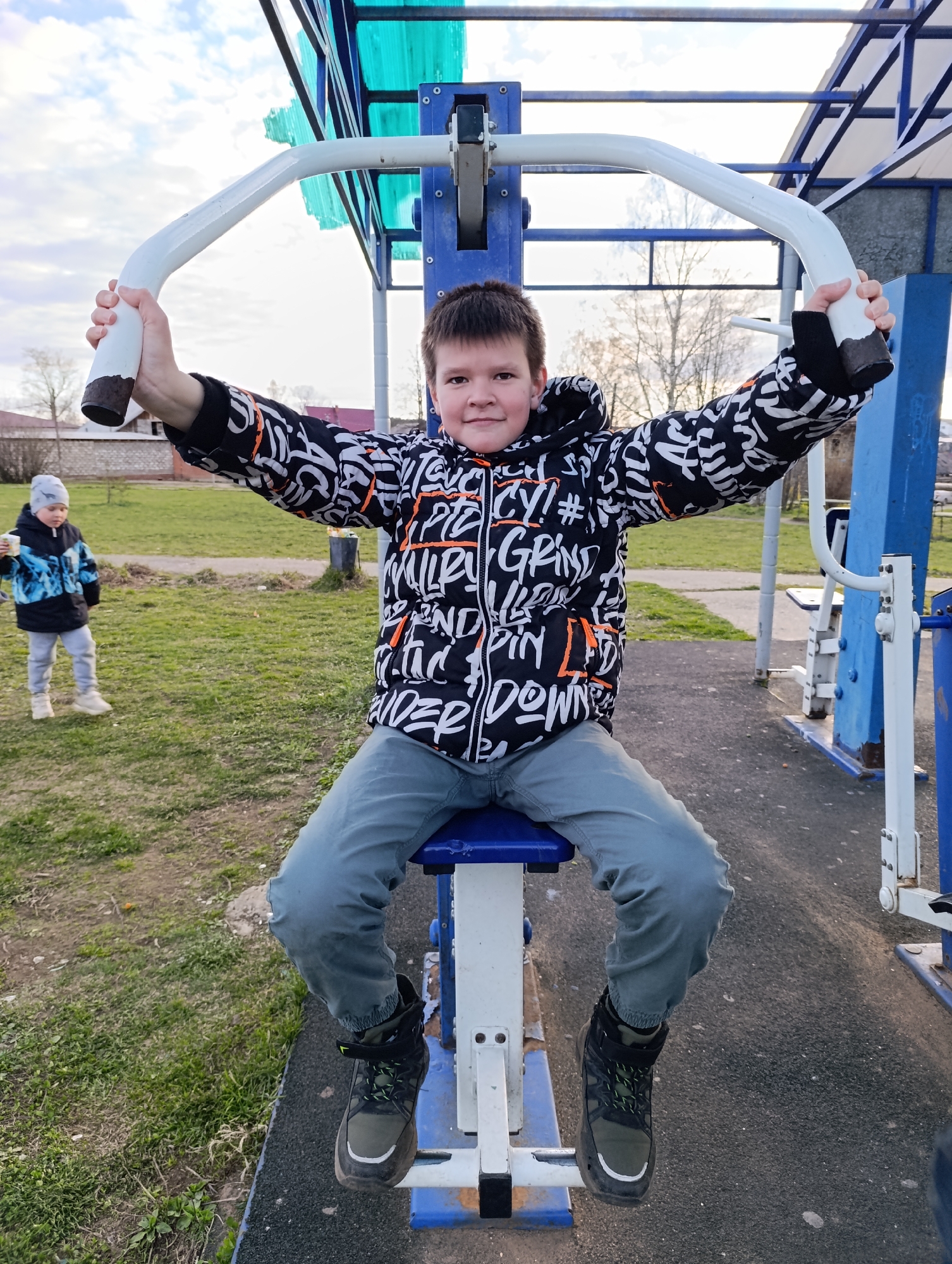 A young boy is sitting on a piece of exercise equipment, gripping the handlebars with outstretched arms, in what appears to be an outdoor exercise area. He is wearing a black, white, and orange hooded jacket with letter designs, grey pants, and dark-colored hiking-style shoes. In the background, slightly out of focus to the left, another child wearing a blue and black jacket is standing on the grass. The exercise equipment is blue and white, and the area features patches of grass and a cloudy sky. The location is Фоки, Russia.
