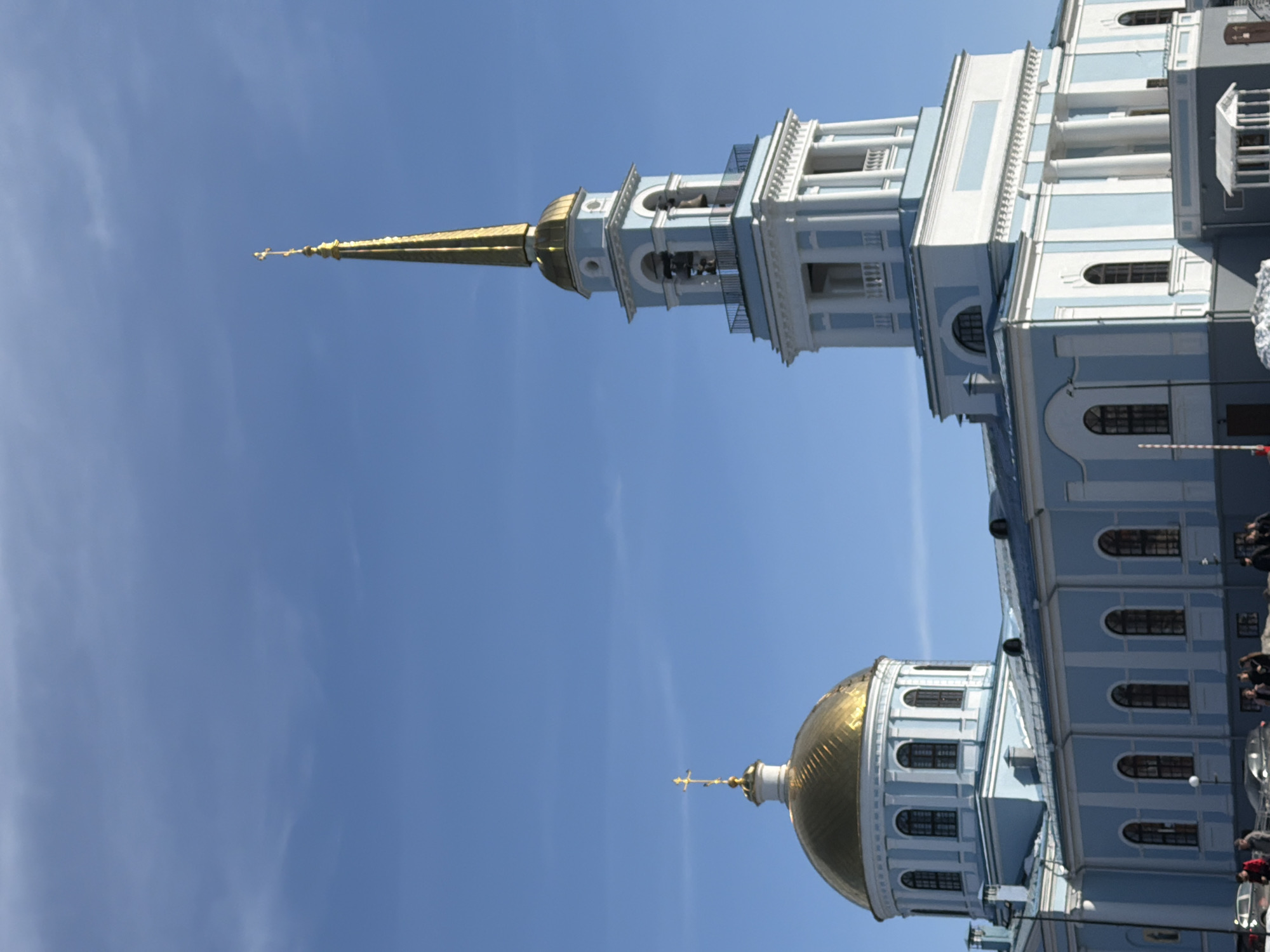 The image depicts the Annunciation Cathedral in Votkinsk, Russia. The cathedral features a blue and white exterior, a golden dome on one end and a tall, gold-tipped spire on the other. There are several windows and architectural details visible. People are standing near the base of the building. The background is a clear, blue sky.