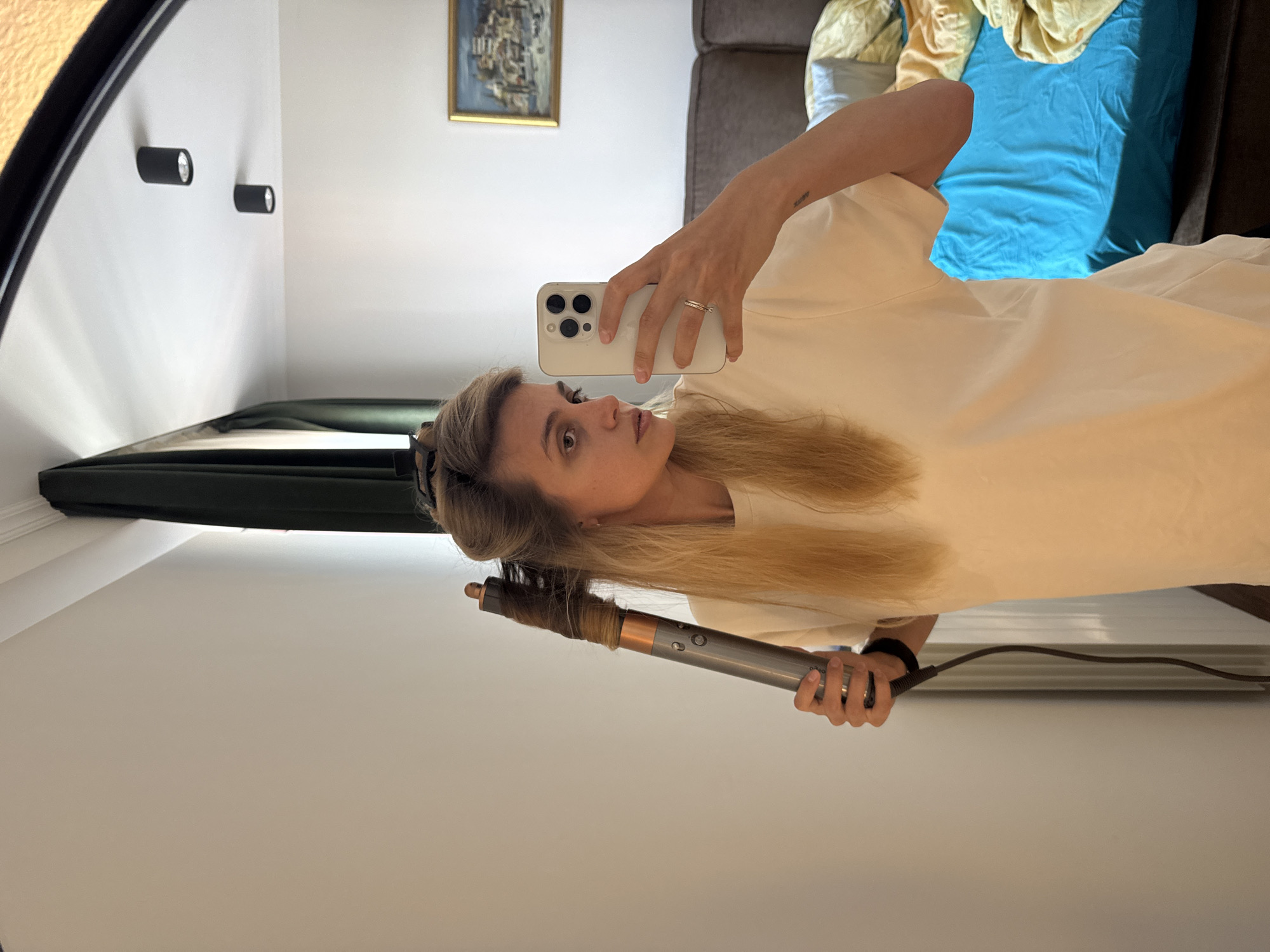 A woman is holding a Dyson Airwrap and a smartphone while posing for a picture in an indoor environment. The background includes wall-mounted lights, a framed picture, a grey sofa, and a bed with blue and yellow linens. The city and state are Unknown.