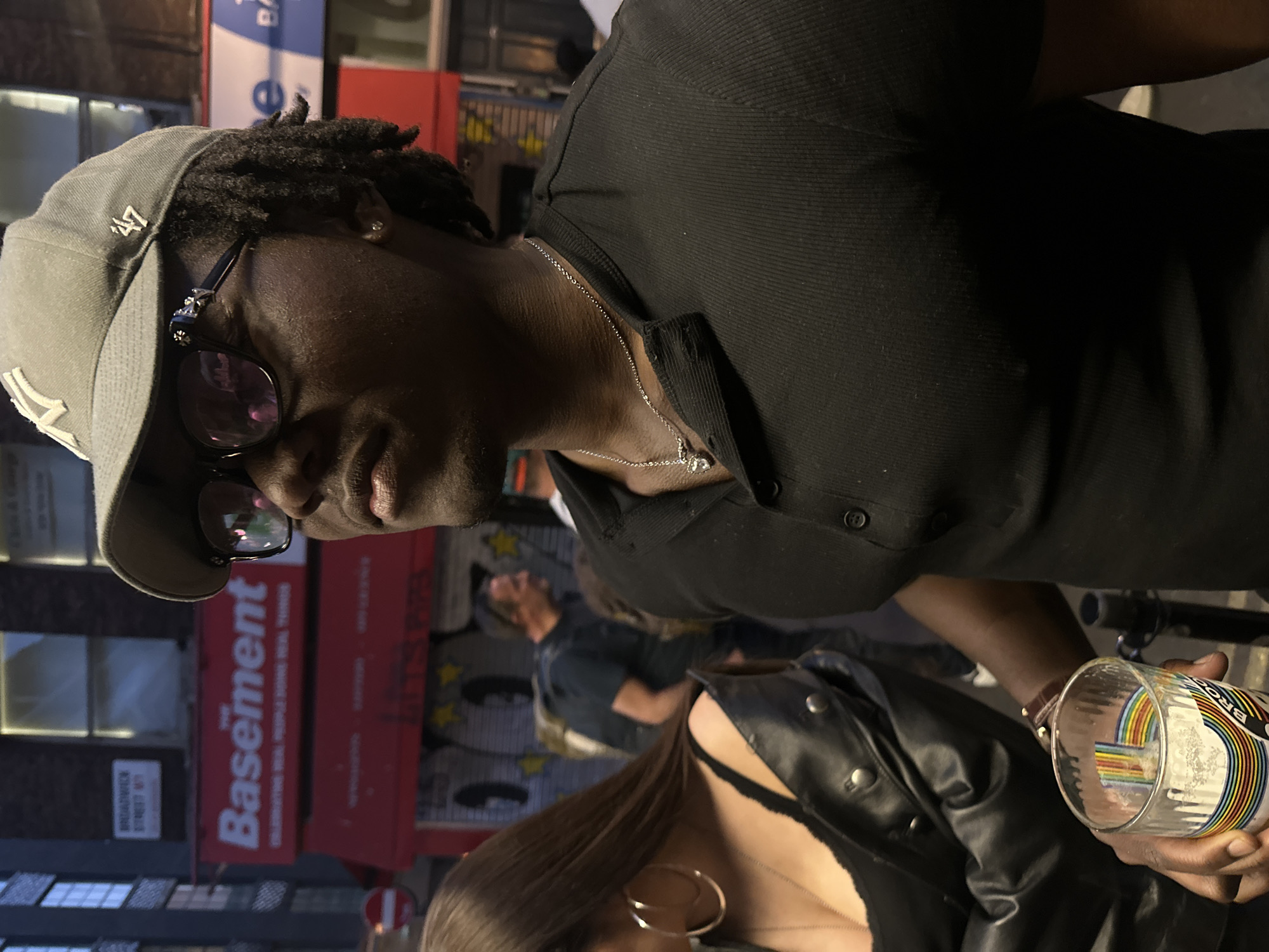 The image depicts an individual wearing a gray baseball cap, dark sunglasses, and a black collared shirt. They are holding a partially empty beer glass decorated with a rainbow motif. A second person, wearing a black leather jacket and a gold hoop earring, is partially visible in the foreground. The background shows a street scene, with a sign that reads "Basement" and "Broadway Street W1", indicating a location in the city of London, United Kingdom. Other individuals can be seen blurred in the background. The overall lighting suggests an evening setting.