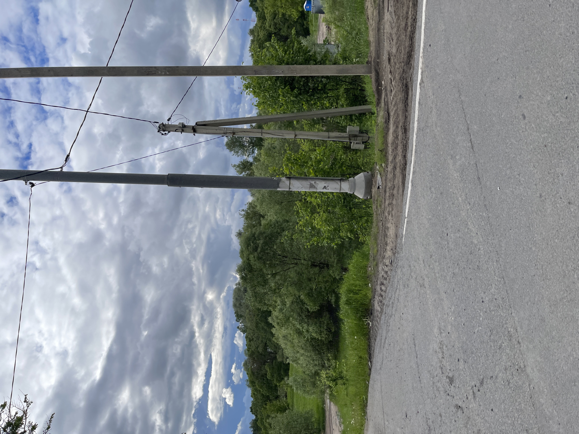 The image presents an oblique view of a roadside scene in Semenovskoe, Russia. On the left side, there are two utility poles, one with visible power lines, set against a backdrop of cloudy blue sky. The poles are adjacent to a strip of green vegetation, beyond which lies a dense line of trees. The right side of the image is dominated by a paved road with a white shoulder line, showing typical wear and tear. The perspective suggests the shot was taken from a low angle, with the camera angled upwards towards the sky. No people are visible in the scene.